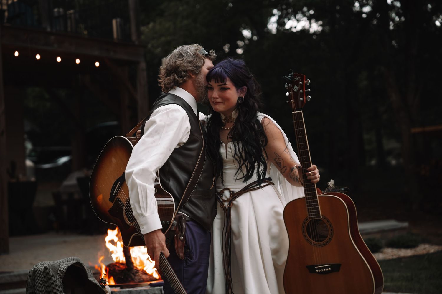 Couple in wedding attire holding acoustic guitars by a campfire at an outdoor evening celebration.