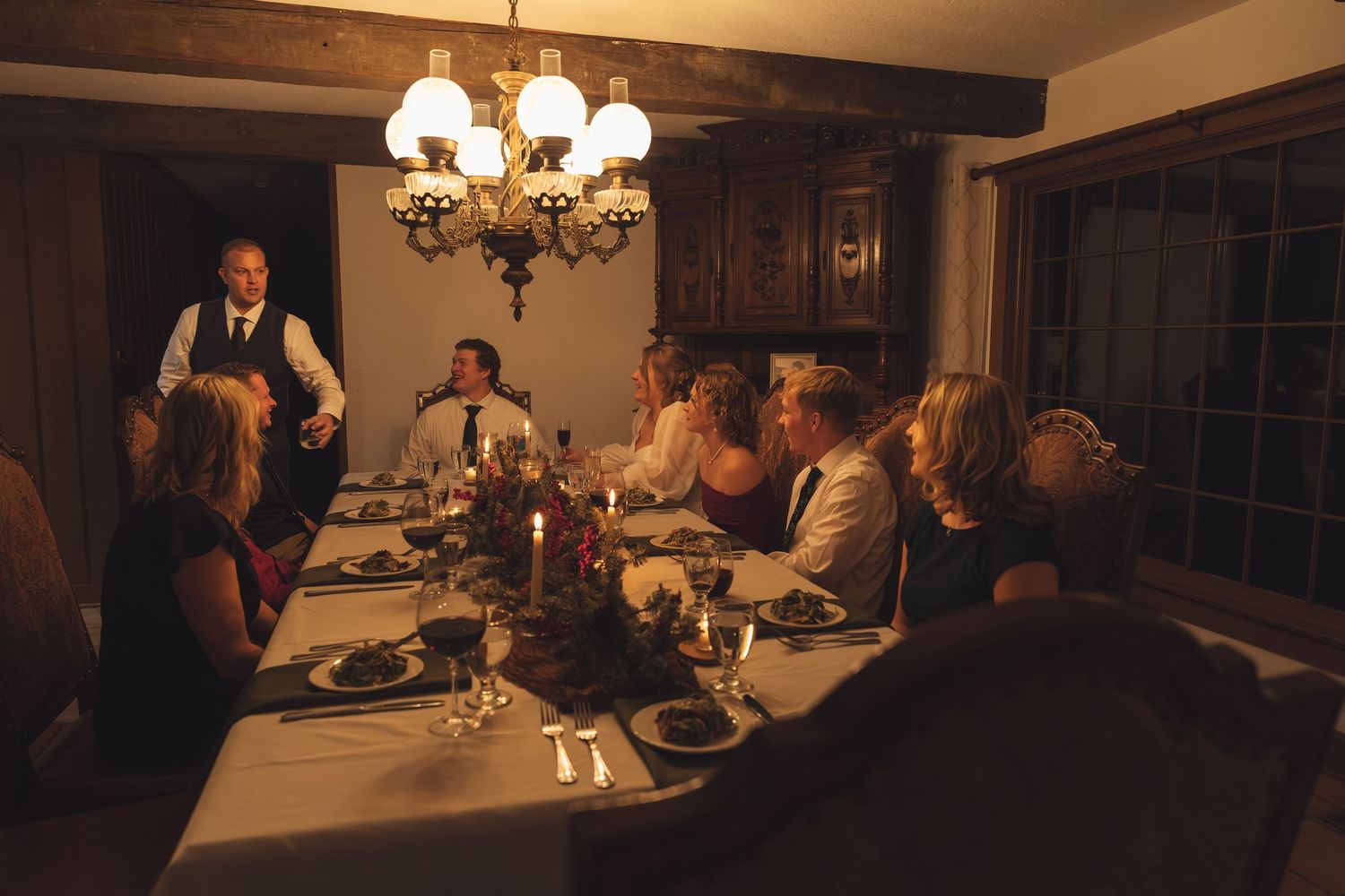 Elegant candlelit dinner party with guests seated at a long dining table under a chandelier in a cozy room.