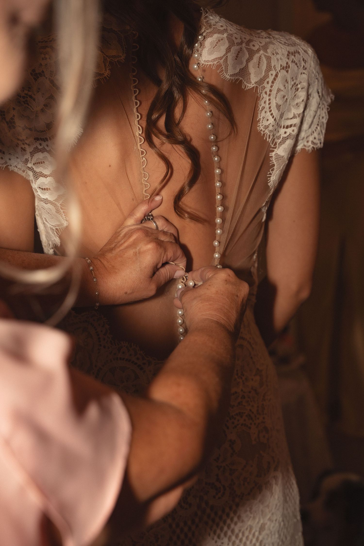 Bride in lace wedding dress being helped with pearl button back closure by bridesmaids during bridal preparation.