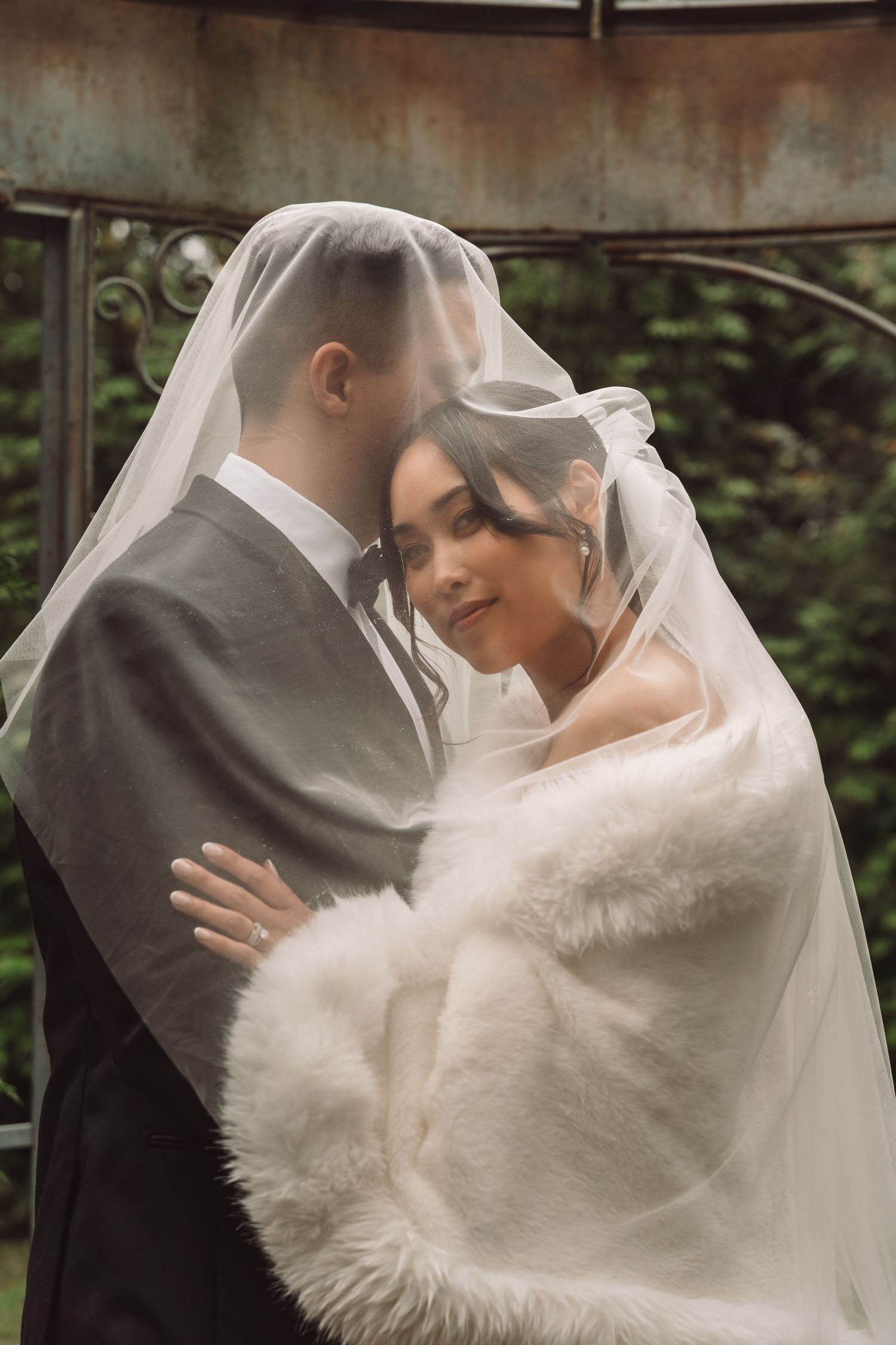 Bride and groom share a romantic moment under a flowing wedding veil outdoors, bride wearing a white fur wrap.