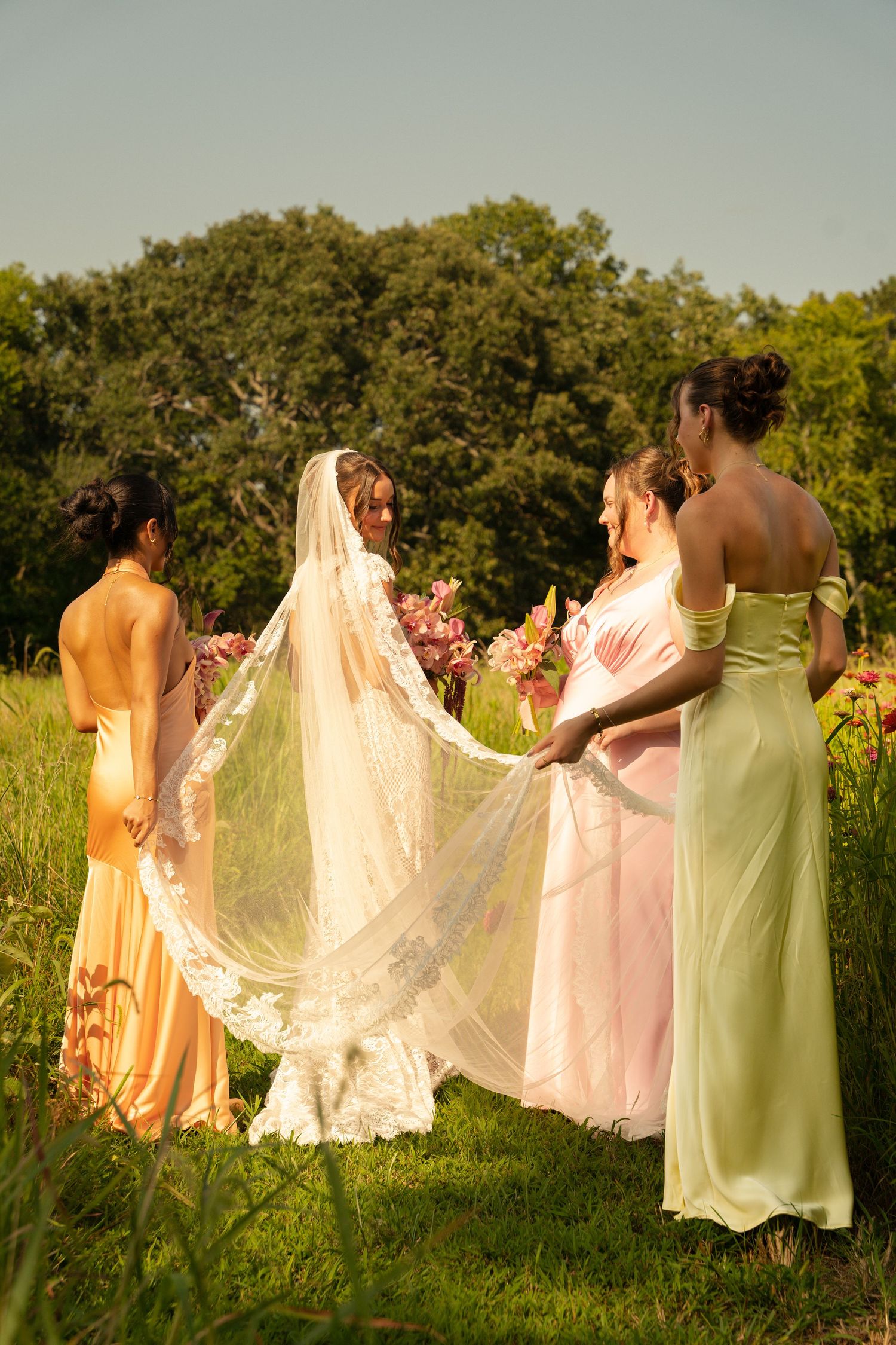 Bride in white gown and lace veil with bridesmaids in pastel dresses standing in a lush green outdoor setting.