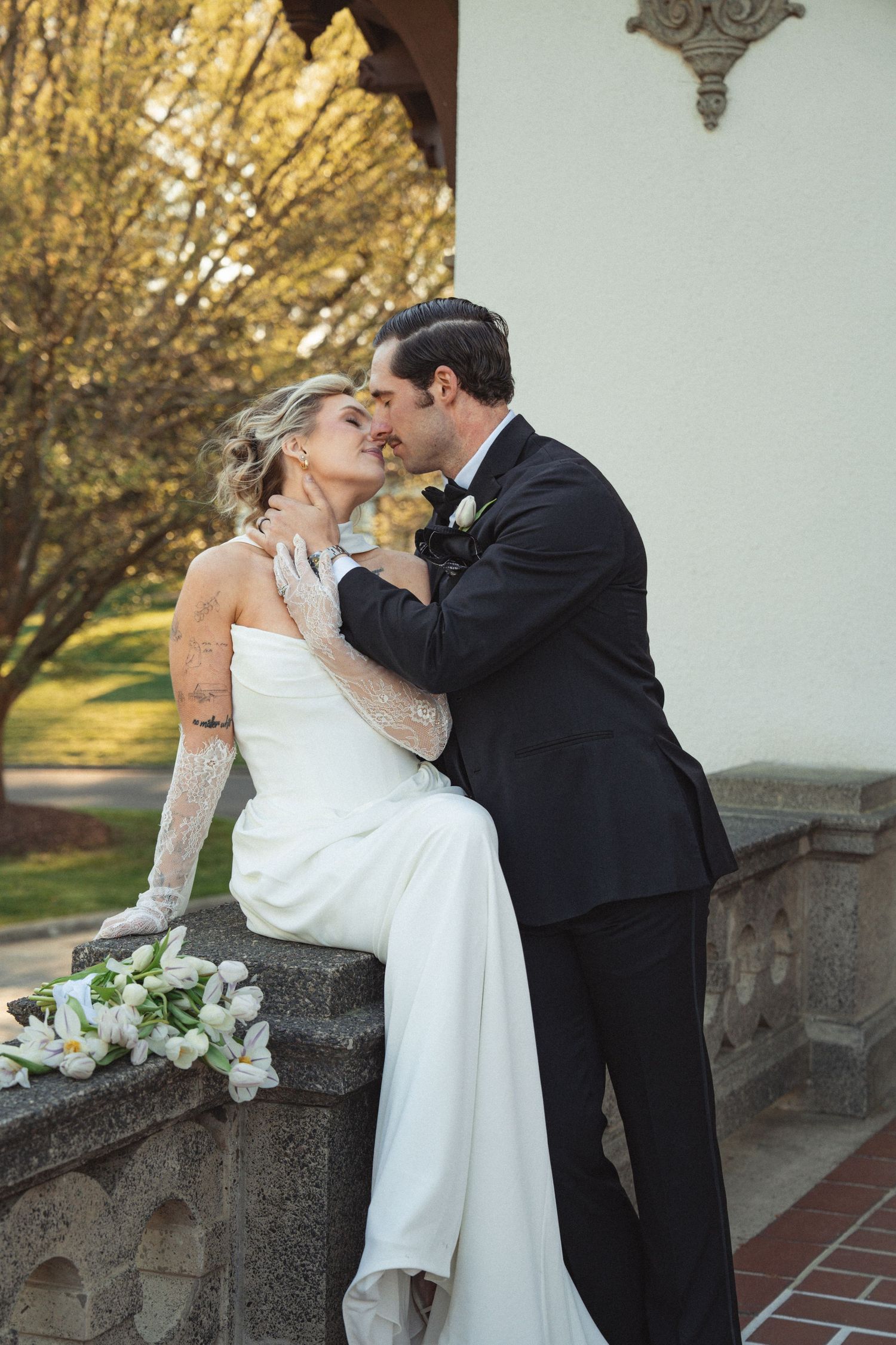 Bride and groom share a romantic almost-kiss on a stone balustrade, bride in white gown with lace gloves holding white florals.