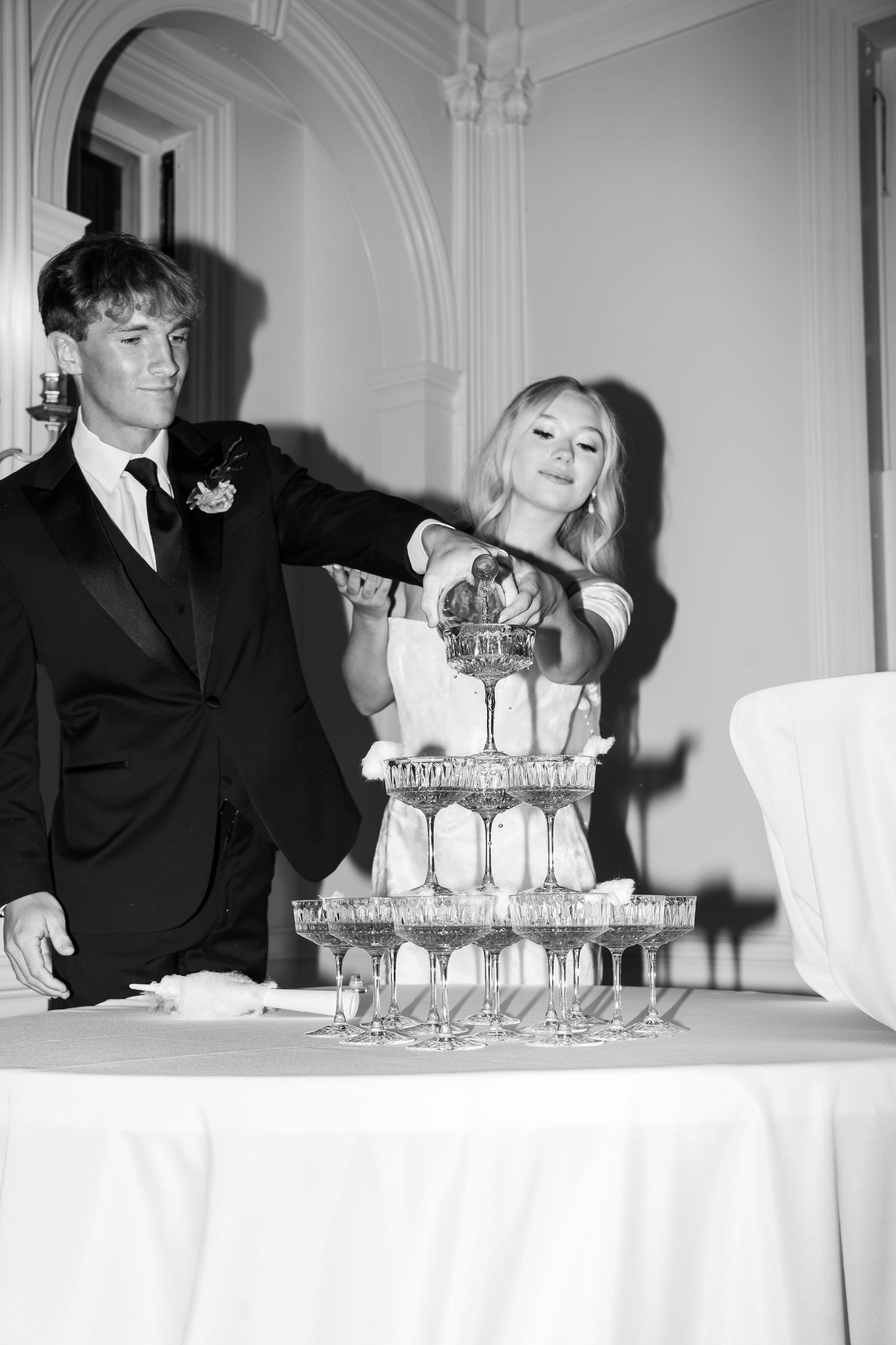 Black and white photo of a bride and groom pouring champagne into a tower of glasses at their wedding reception.