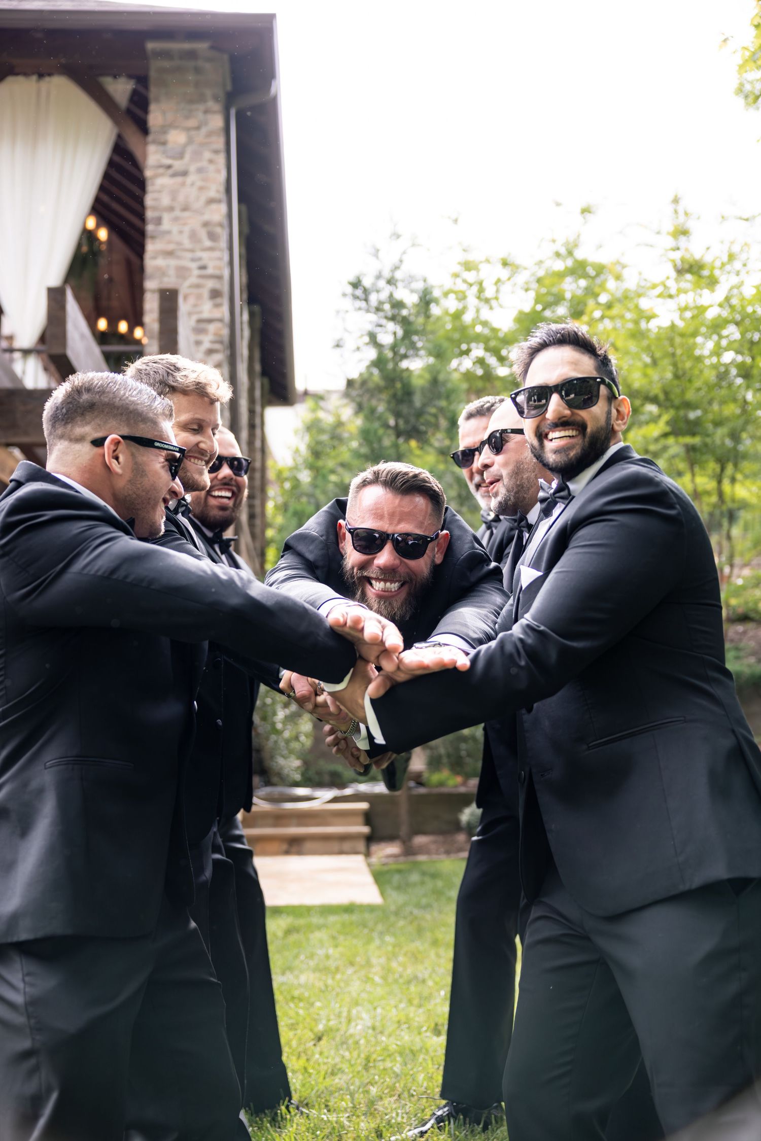 Six groomsmen in black suits and sunglasses stack hands together outdoors at a wedding venue.