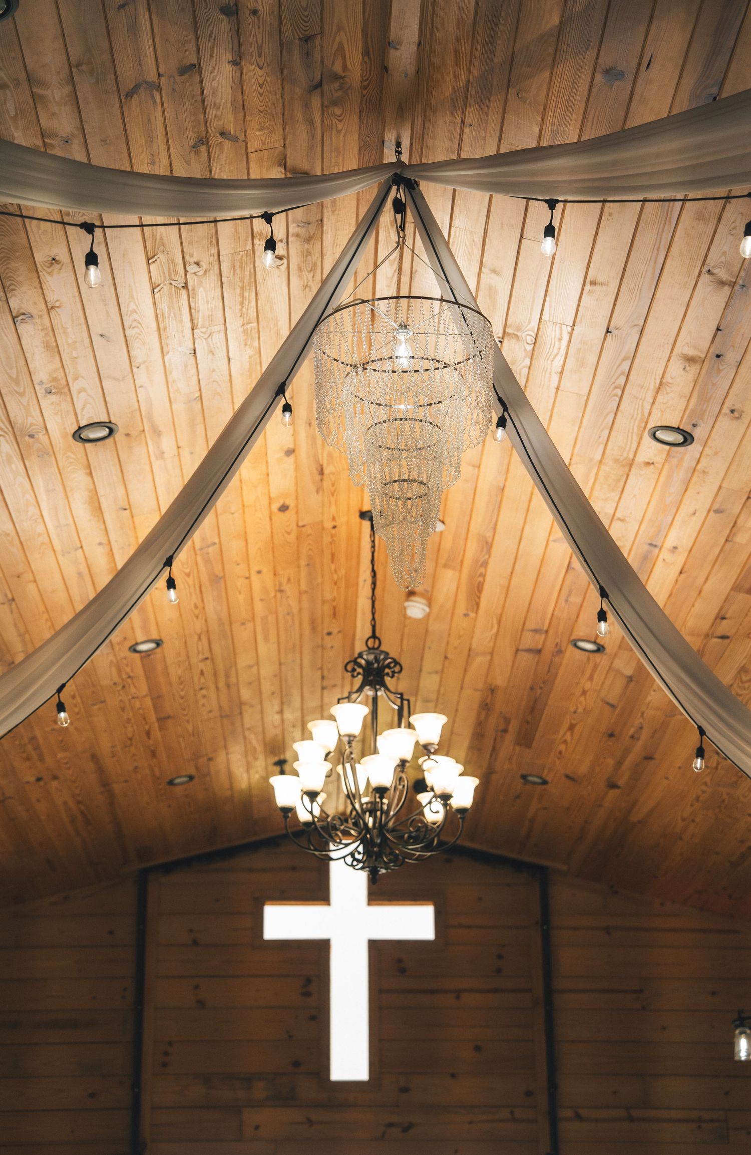 Rustic wooden chapel interior with elegant chandelier, draped fabric ceiling, string lights, and illuminated cross at the altar.