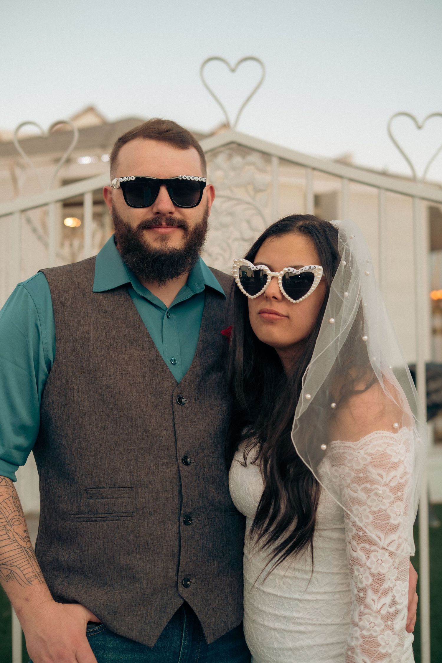 Bride and groom wearing stylish sunglasses pose in front of heart-shaped decor at their outdoor wedding ceremony.