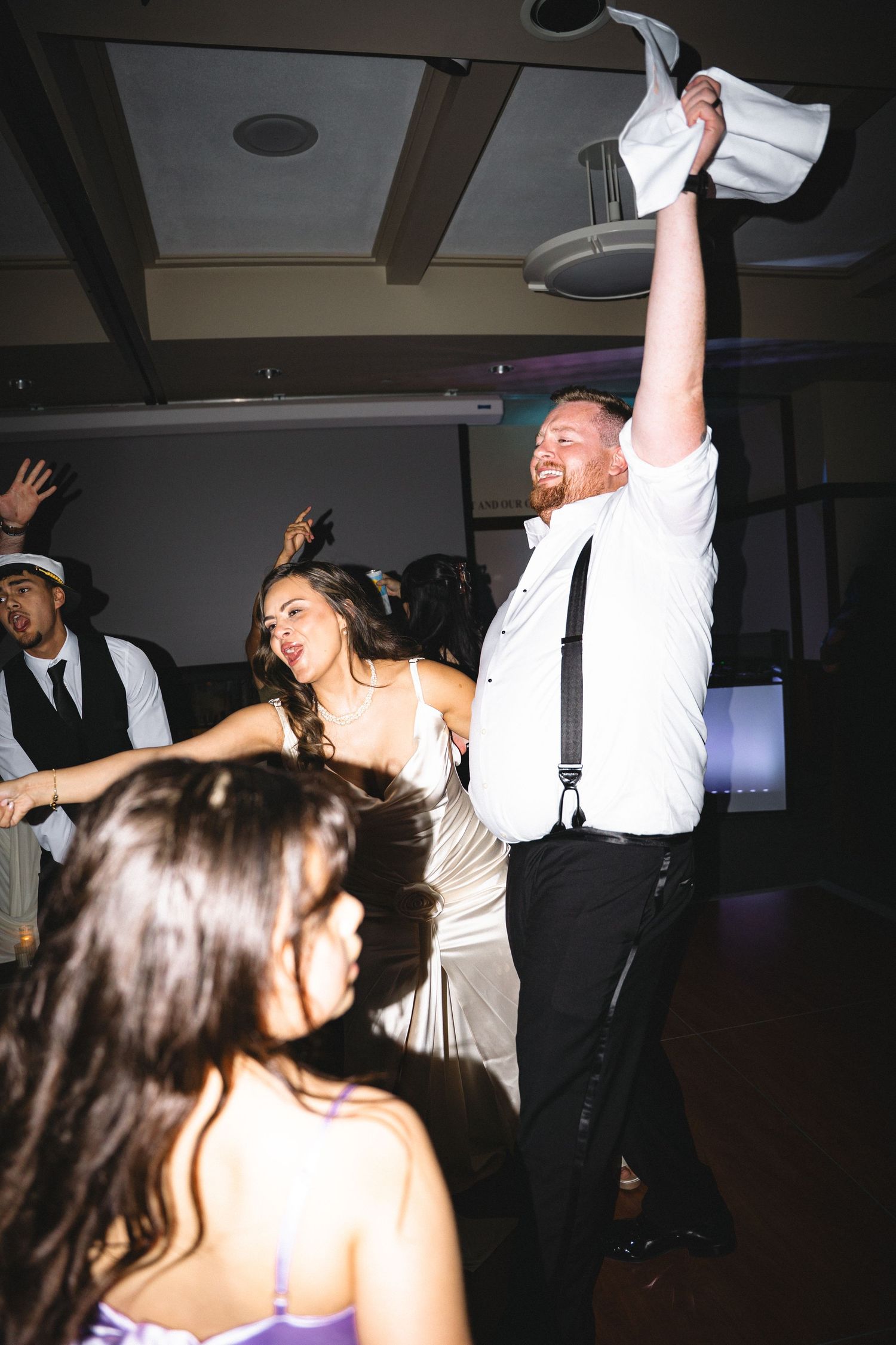 Bride and groom dancing joyfully at wedding reception, groom raising white hat in celebration.
