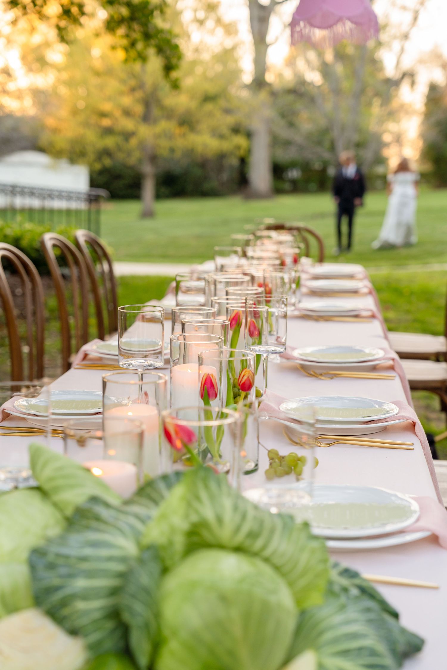 Elegant outdoor wedding reception table set with tulip centerpieces, candles, and cabbage leaves on a lush green lawn.