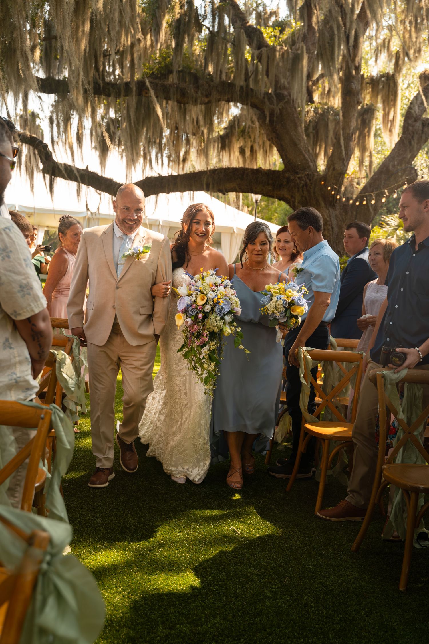 Bride and groom walk down outdoor aisle under Spanish moss tree, flanked by guests and bridesmaid holding colorful bouquet.