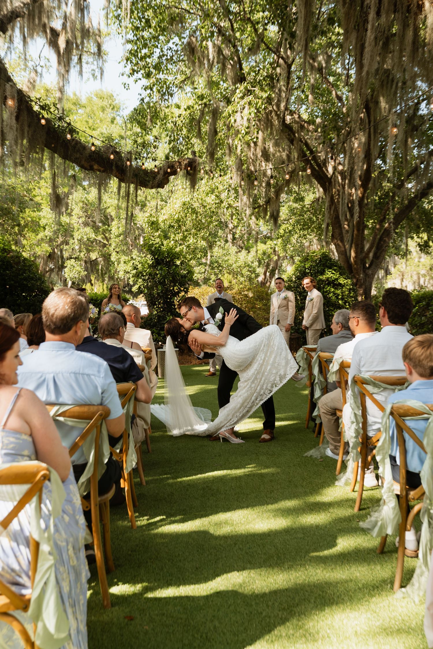 Newlyweds share a romantic dip kiss walking down outdoor wedding aisle lined with guests under moss-draped oak trees.
