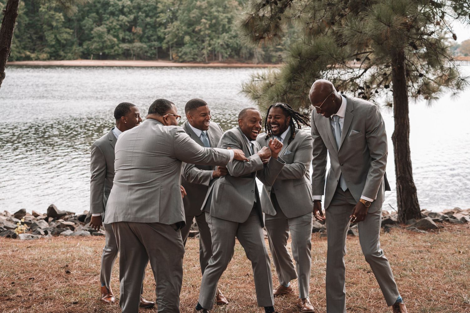 Groomsmen in gray suits laughing and celebrating together near a lakeside during a wedding.