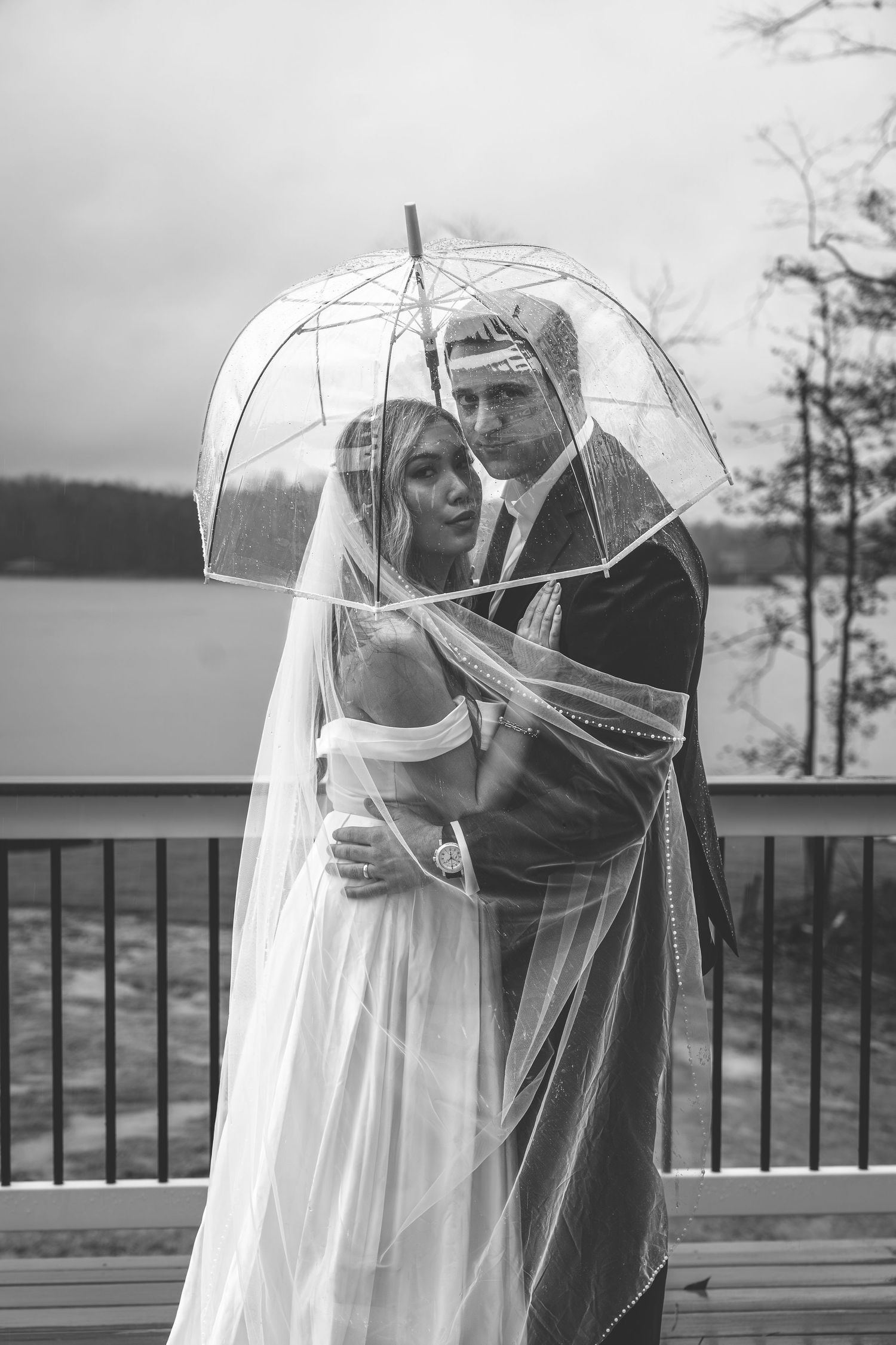 Black and white photo of bride and groom embracing under a clear umbrella outdoors on a rainy wedding day.