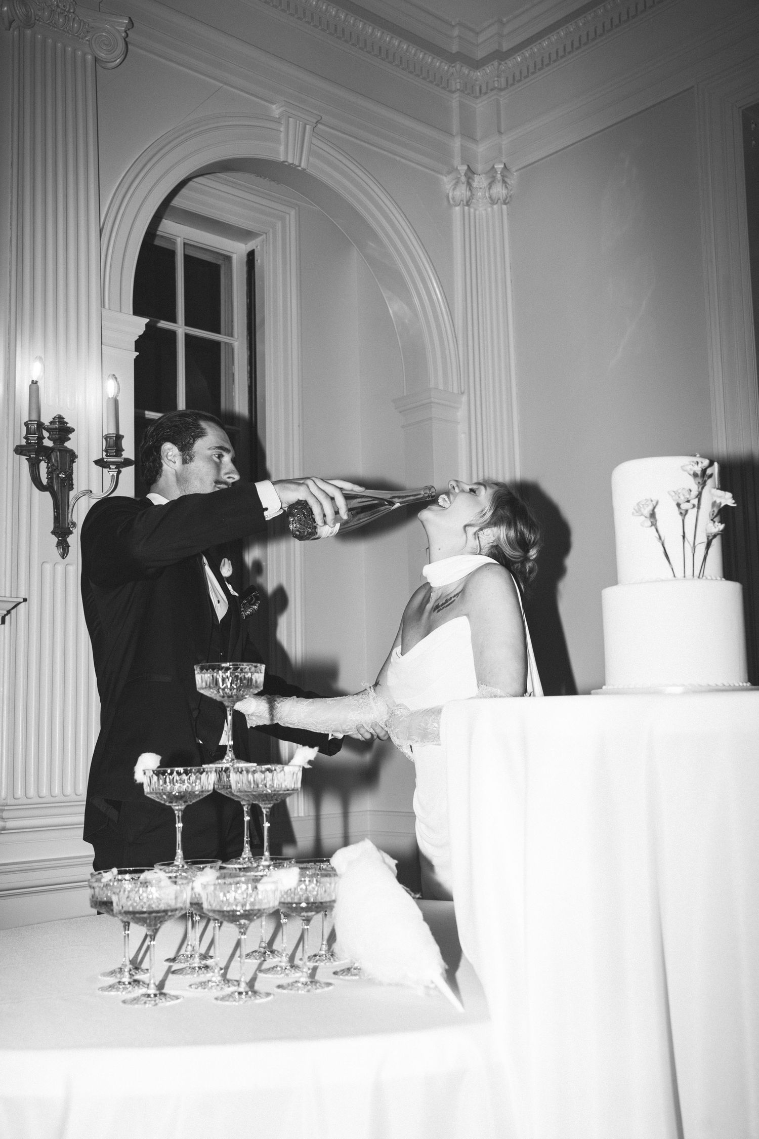 Bride and groom celebrate at wedding reception with champagne tower, groom pouring bubbly into bride's mouth.