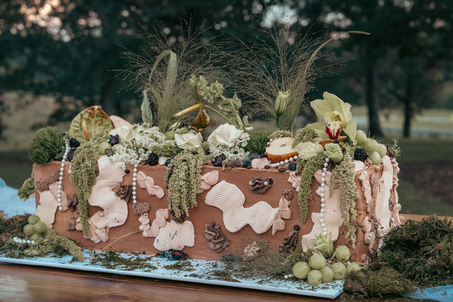 Elaborate floral cake with greenery, feathers, pearls, and blooms on a rustic outdoor table setting.