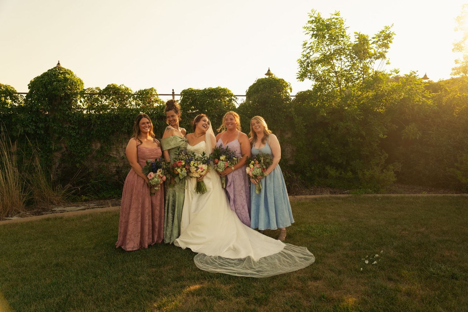 Bride in white gown with five bridesmaids in colorful pastel dresses holding bouquets in a sunlit garden.
