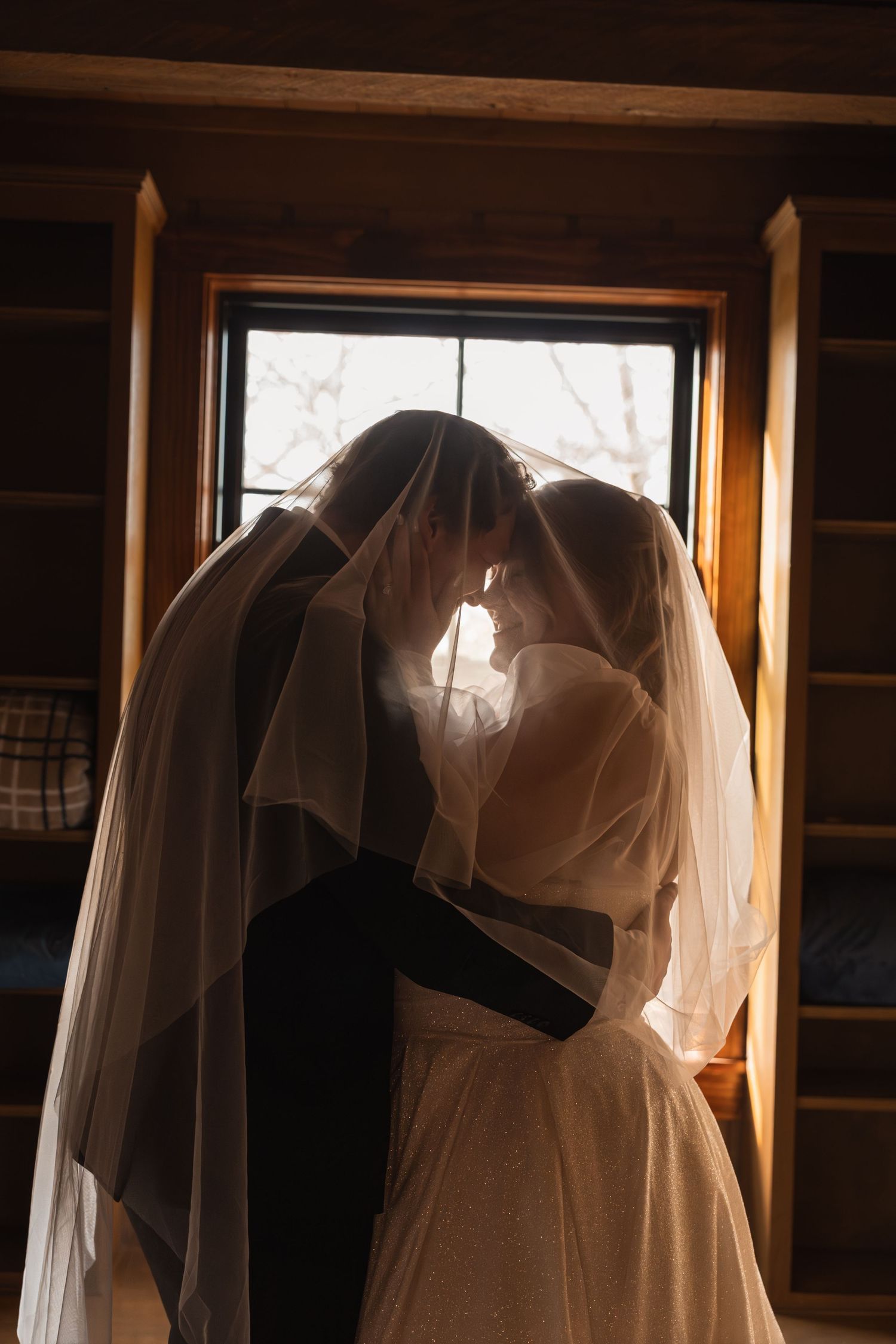 Bride and groom sharing romantic moment under wedding veil, backlit by window in rustic wooden cabin.