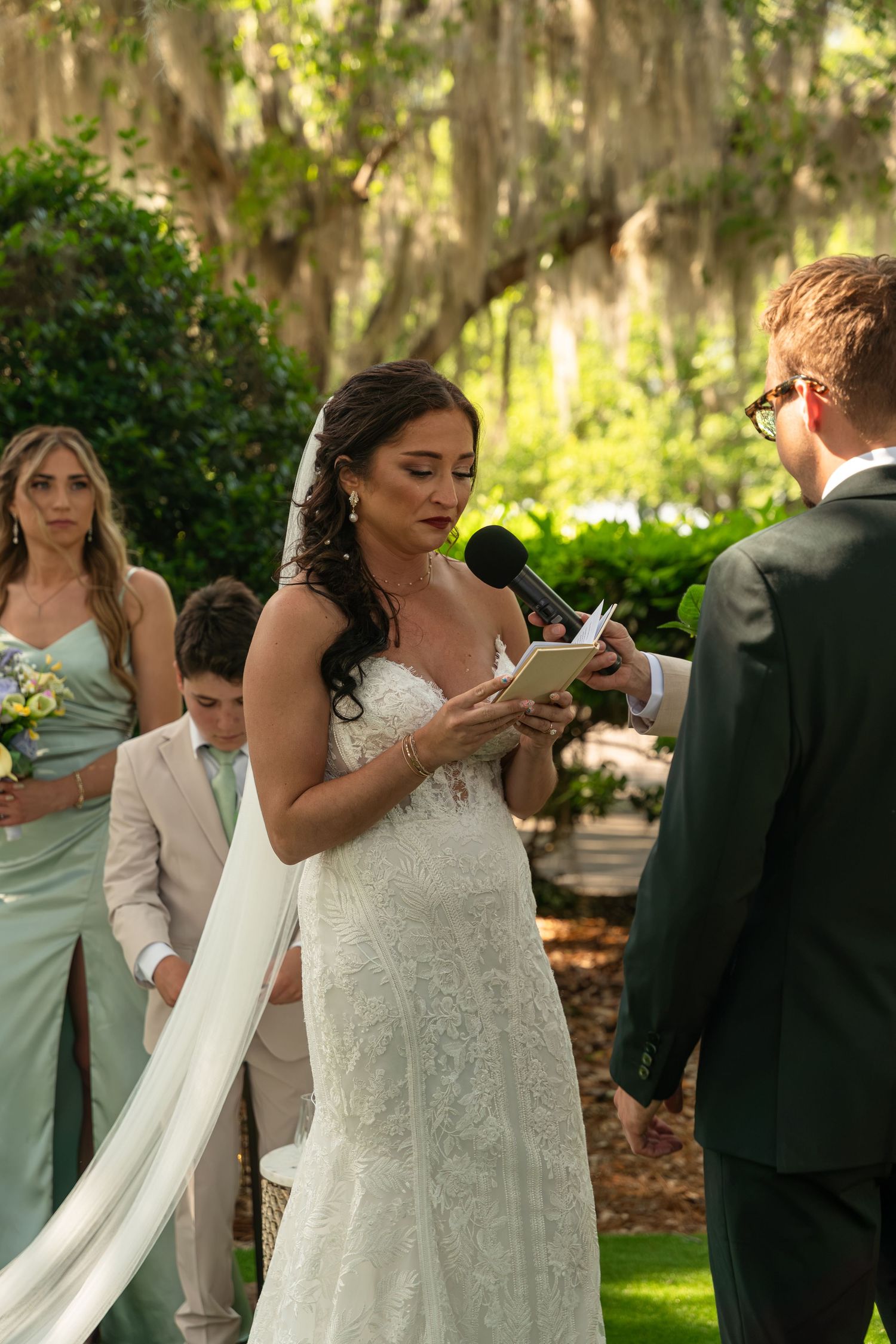 Bride reading wedding vows from a book during outdoor ceremony, wearing lace gown, with bridesmaids and guests behind her.