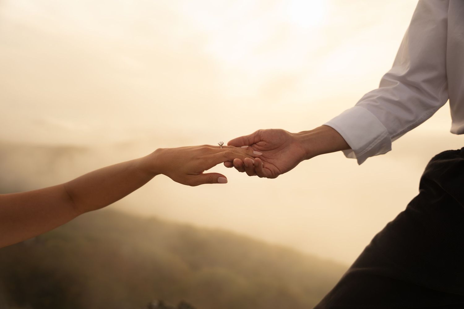 Two people reaching out and holding hands against a soft, hazy golden sky with misty hills in the background.