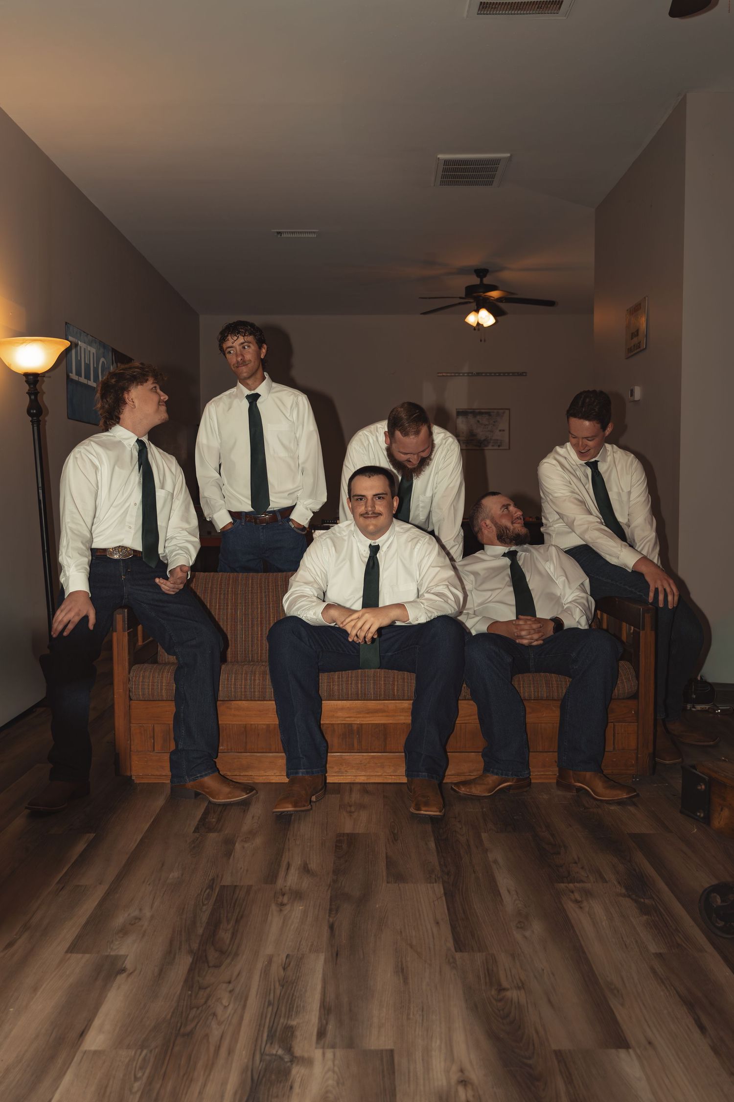 Seven men in white shirts and dark ties pose together on a wooden couch in a warmly lit living room.