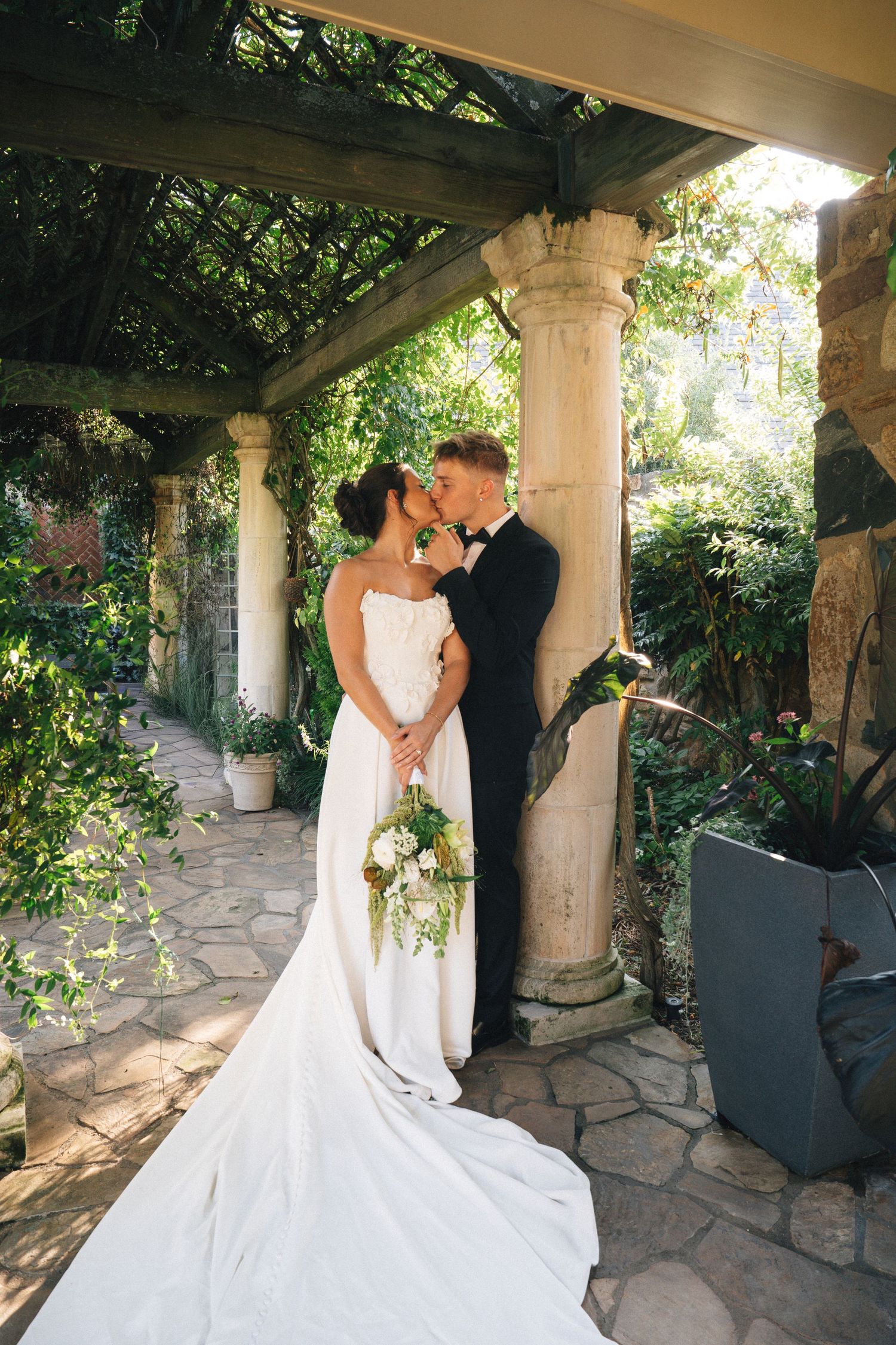 Bride and groom share a romantic moment under a vine-covered pergola with stone columns in a lush garden setting.