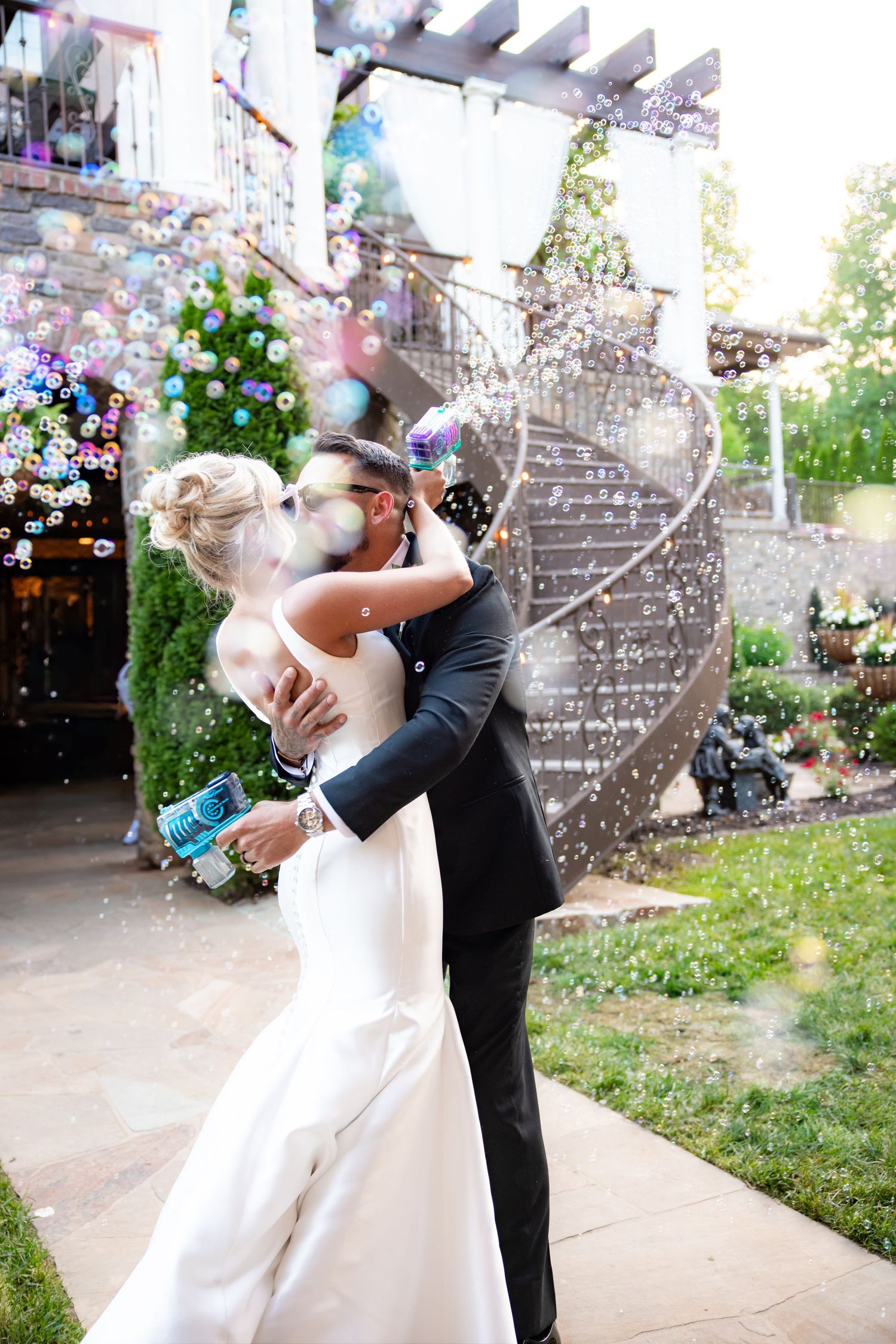 Newlywed couple sharing first kiss surrounded by bubbles outdoors at elegant wedding venue.