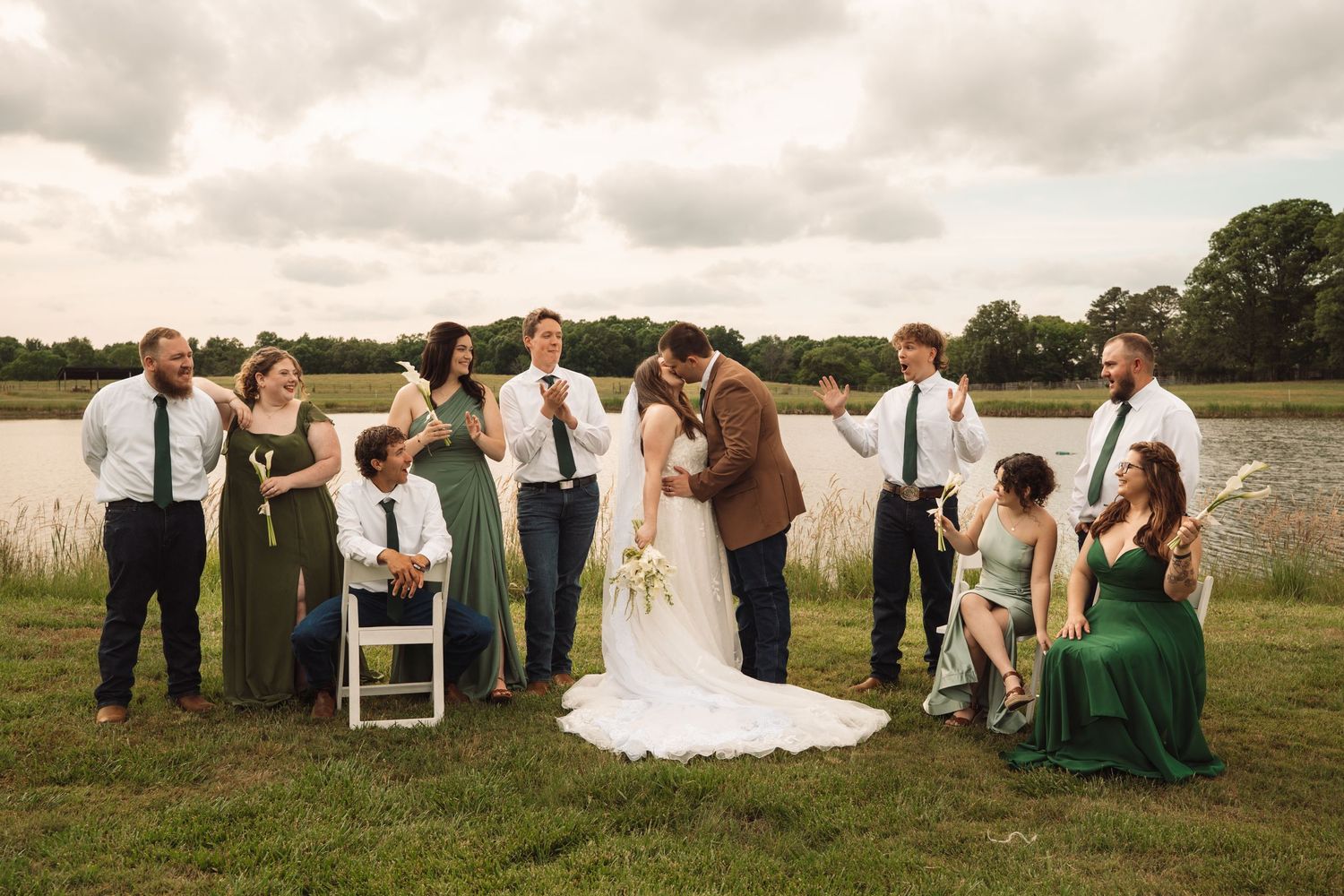 Wedding party celebrating outdoors near a pond, bride and groom kissing while bridal party cheers in green dresses and ties.
