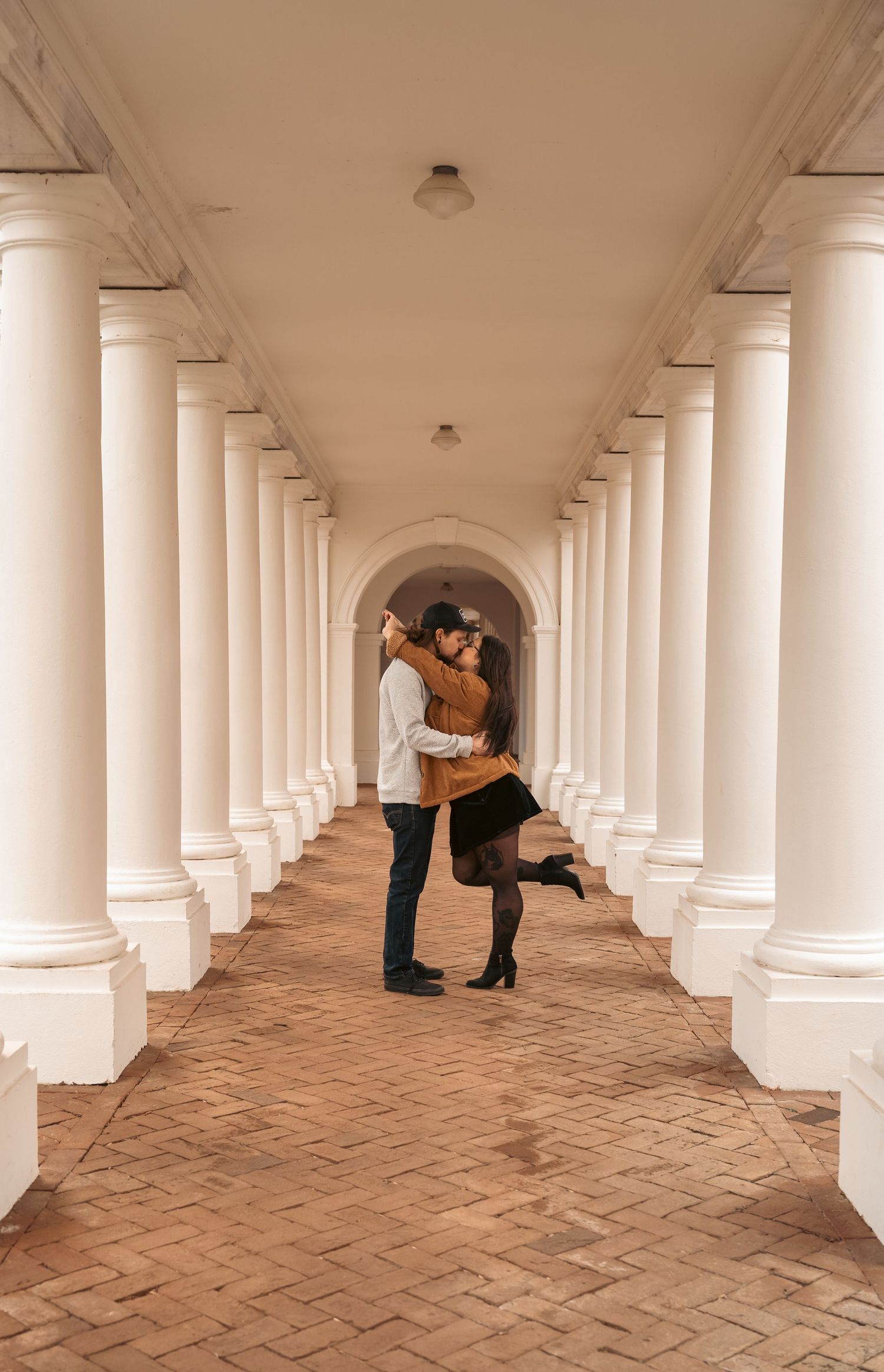 Romantic couple embracing and kissing in a stunning white columned archway corridor with brick flooring.