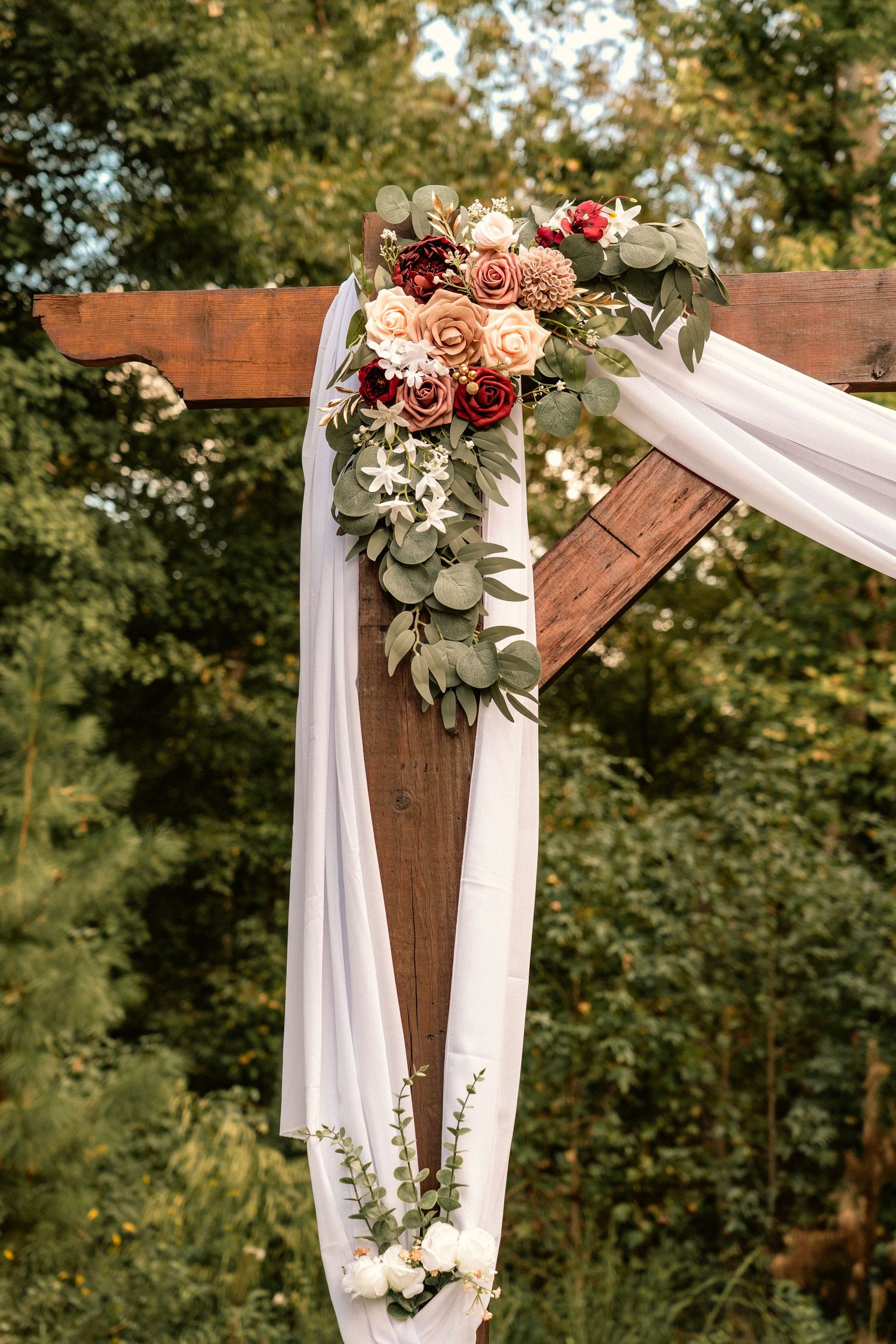 Rustic wooden wedding arch adorned with white draping and a floral arrangement of peach, burgundy roses and eucalyptus outdoors.