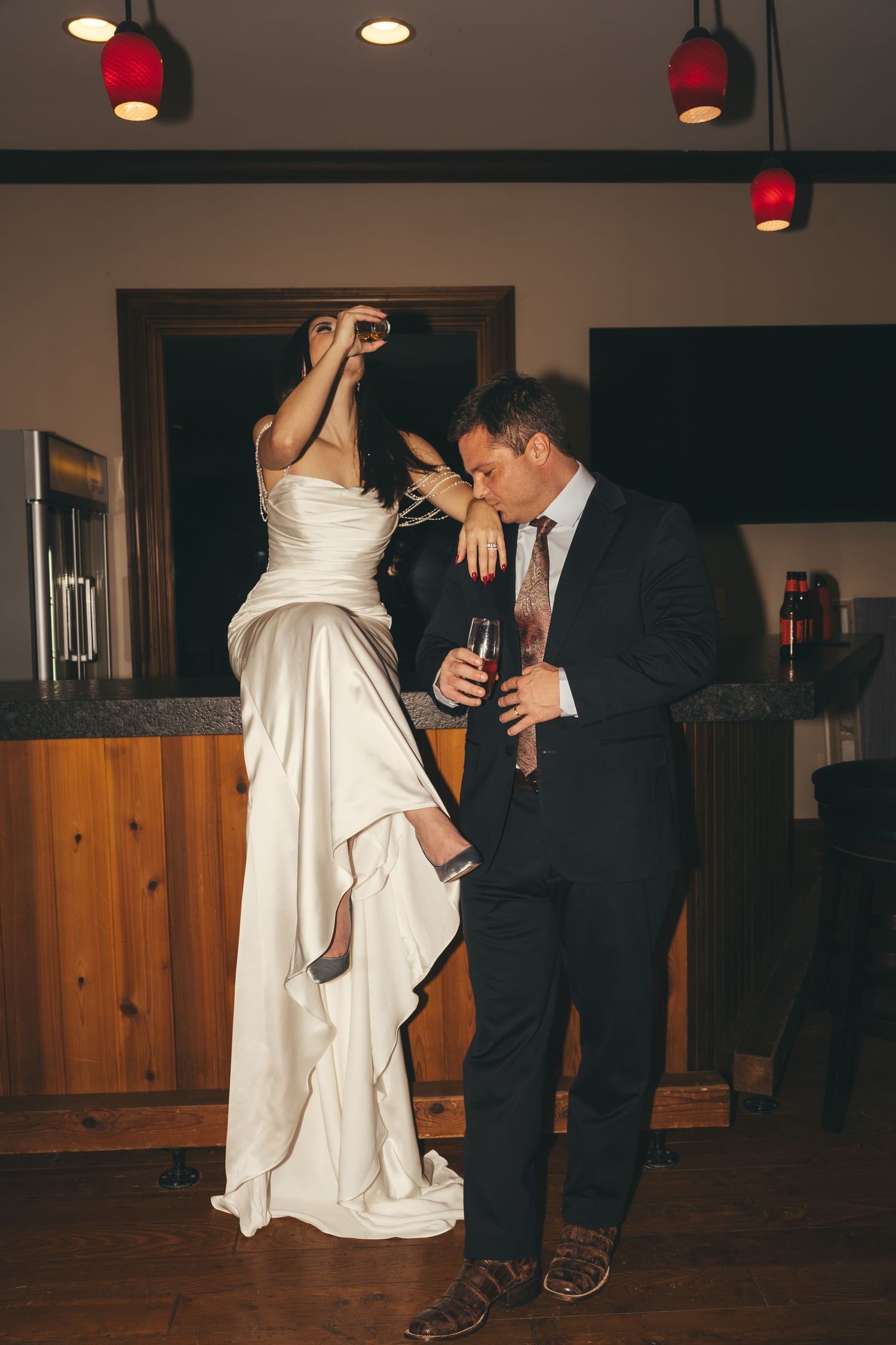 Bride in a satin wedding gown drinks from a glass while leaning on a groom in a suit holding a drink at a rustic wooden bar.