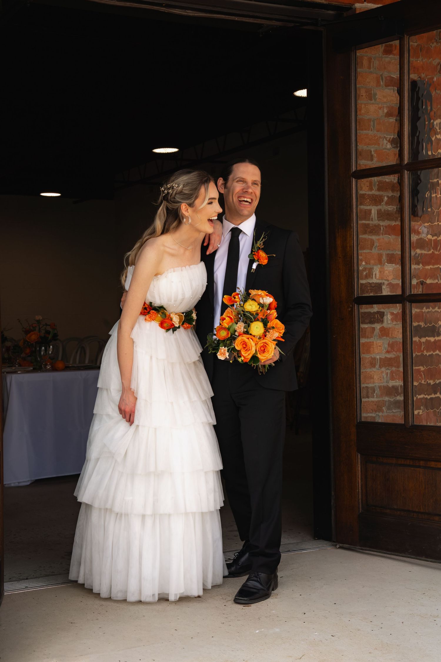 Smiling bride and groom exit through large wooden doors, bride in tiered white gown holding vibrant orange wedding bouquet.