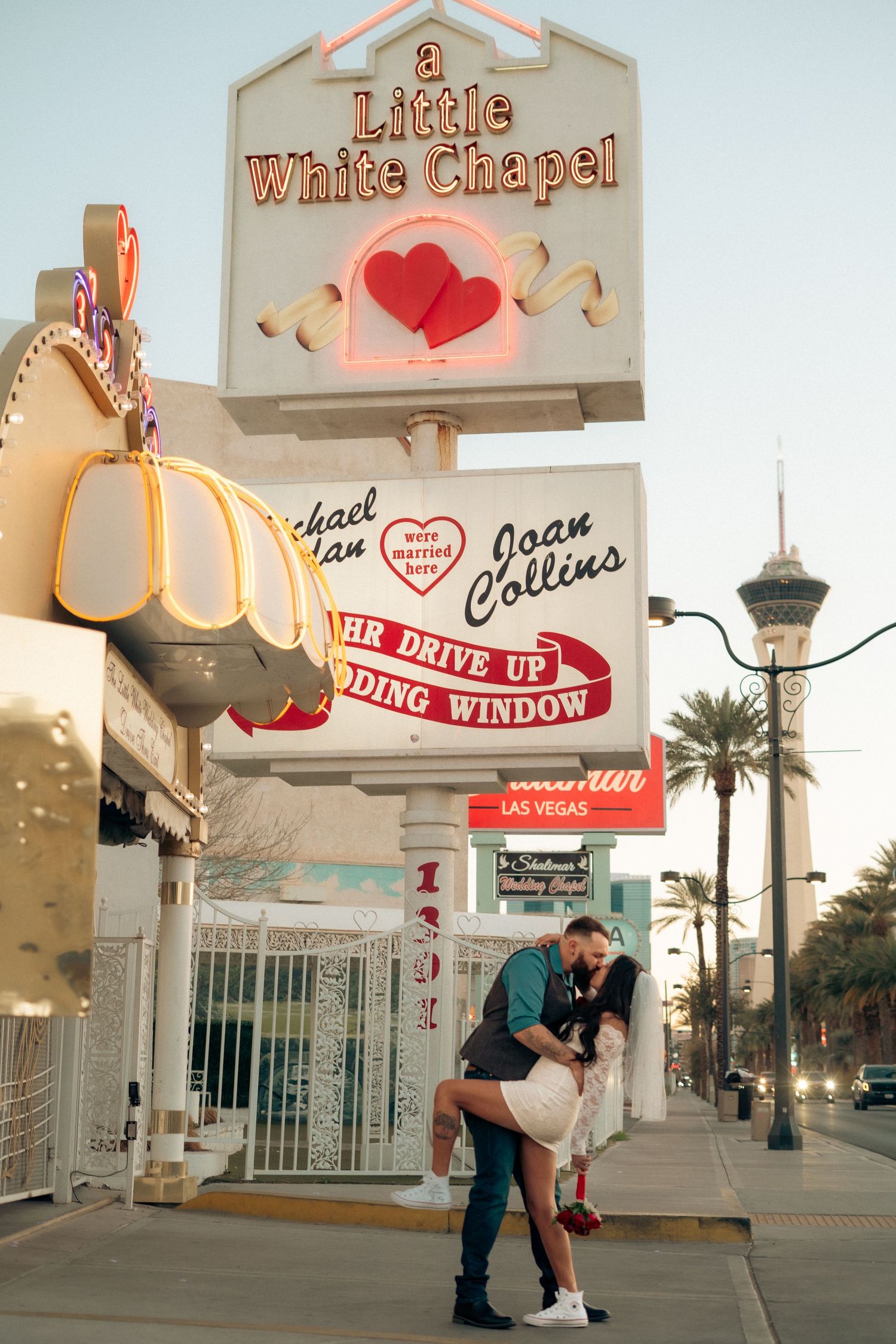 Couple embracing in front of A Little White Chapel wedding sign in Las Vegas, with the Stratosphere tower visible.