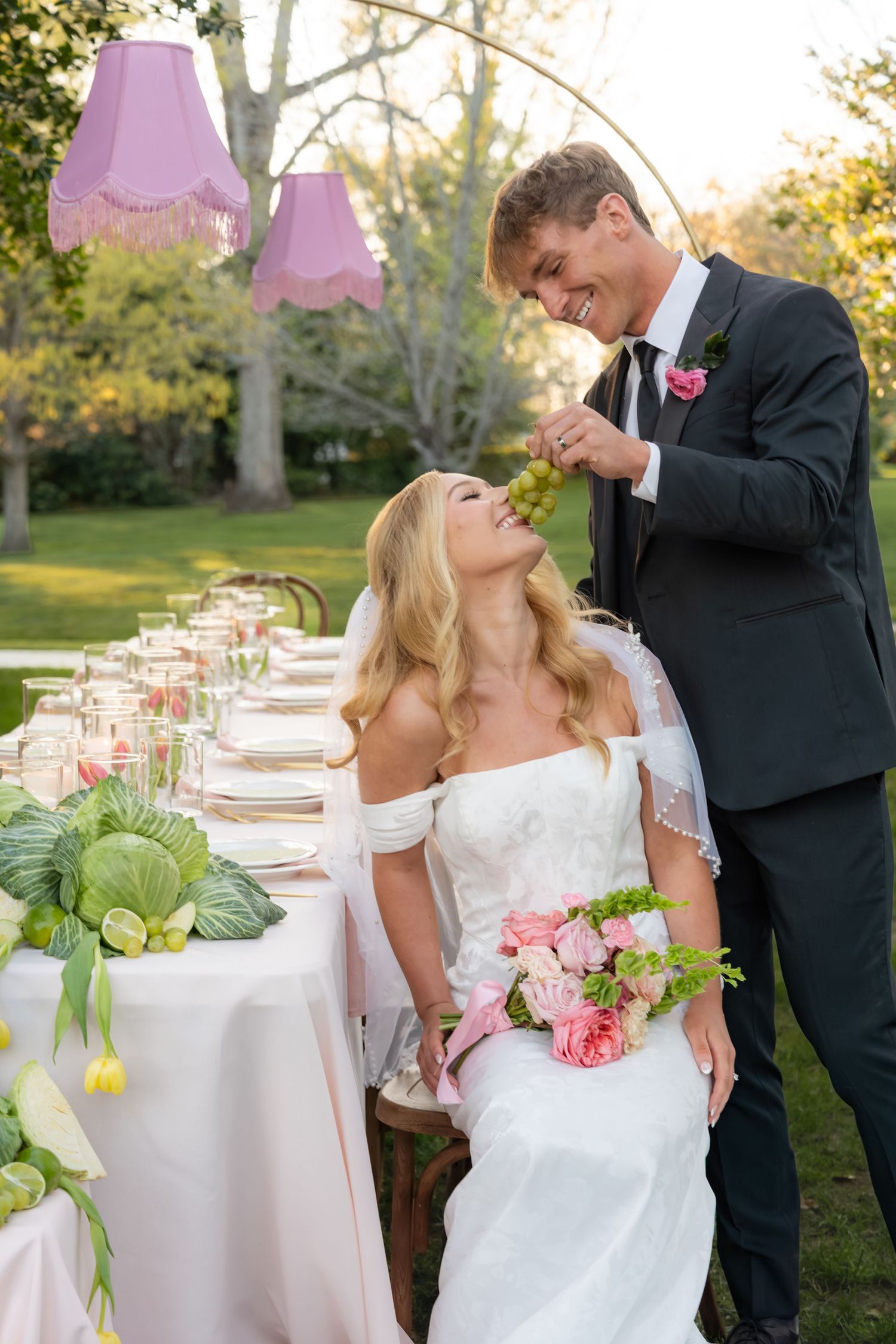 A smiling groom feeds his seated bride a grape at a beautifully decorated outdoor wedding reception table.