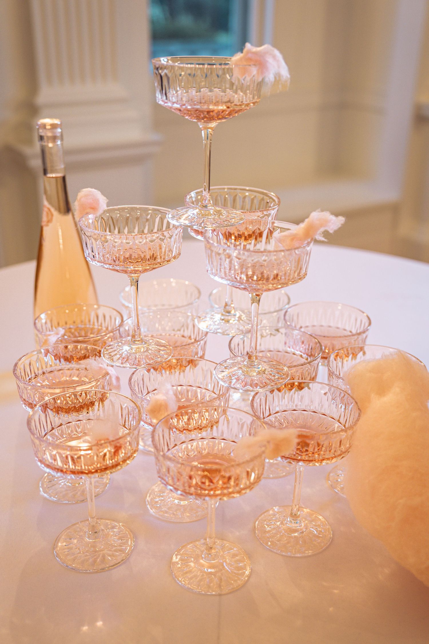 Crystal champagne tower with rose wine and pink feather decorations on white table at elegant celebration.