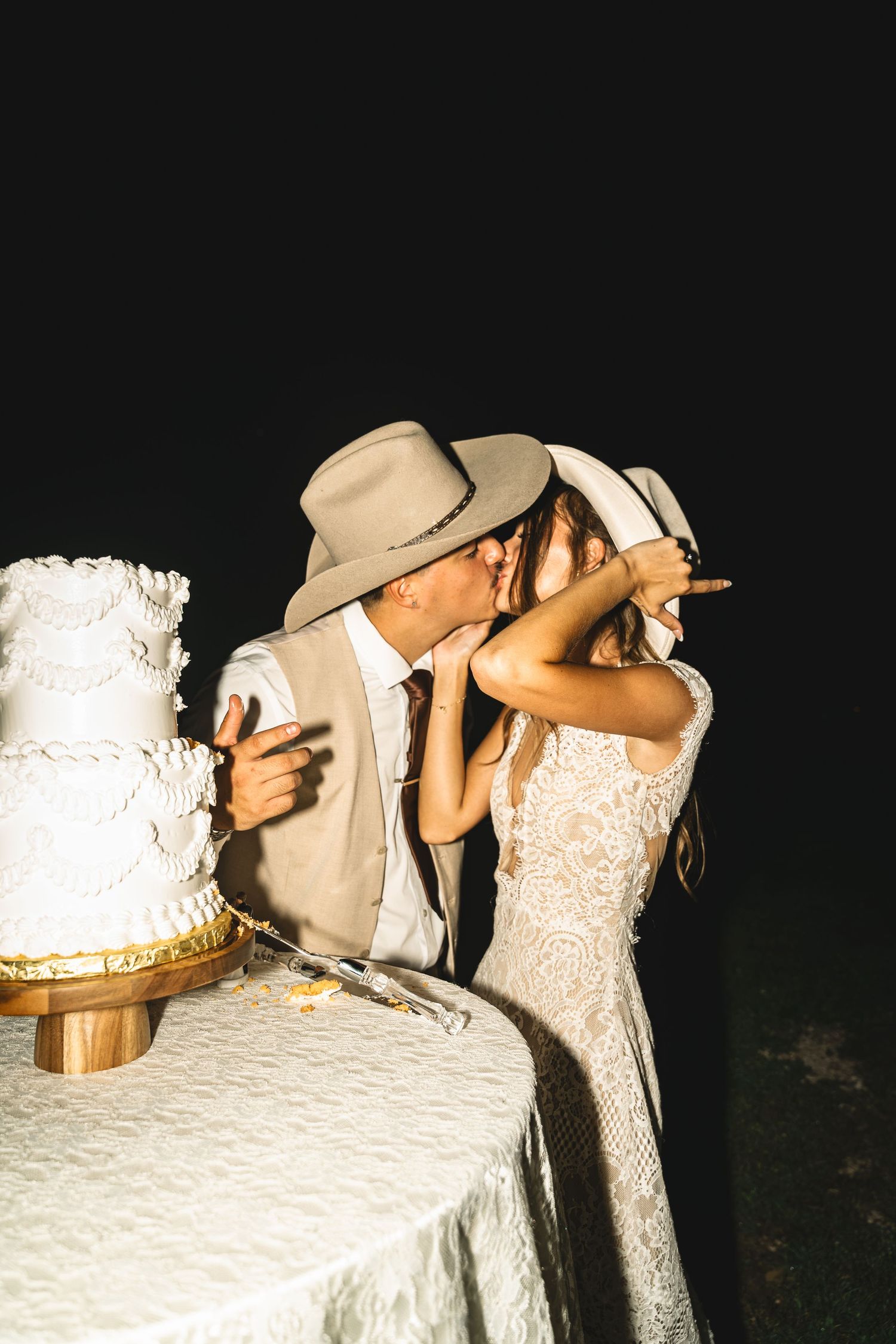 Bride and groom in cowboy hats share a kiss beside their white tiered wedding cake at an outdoor night reception.