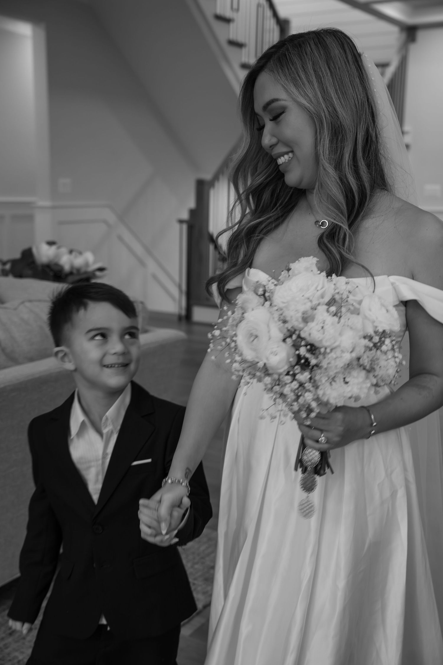 Smiling bride in white gown holds bouquet while young boy in suit holds her hand indoors.