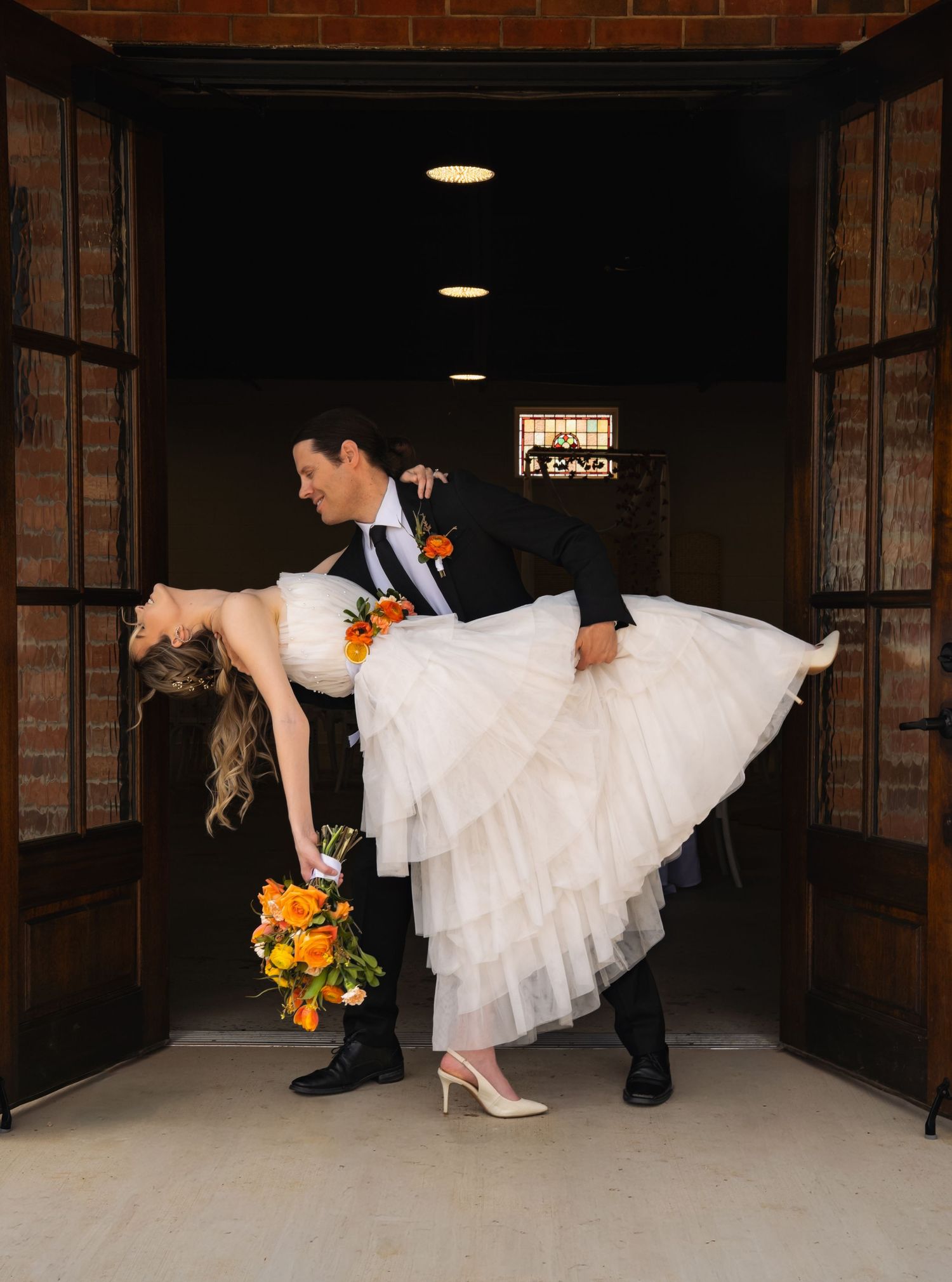 Groom lifts bride in white tiered dress at wooden doors, holding orange bouquet.
