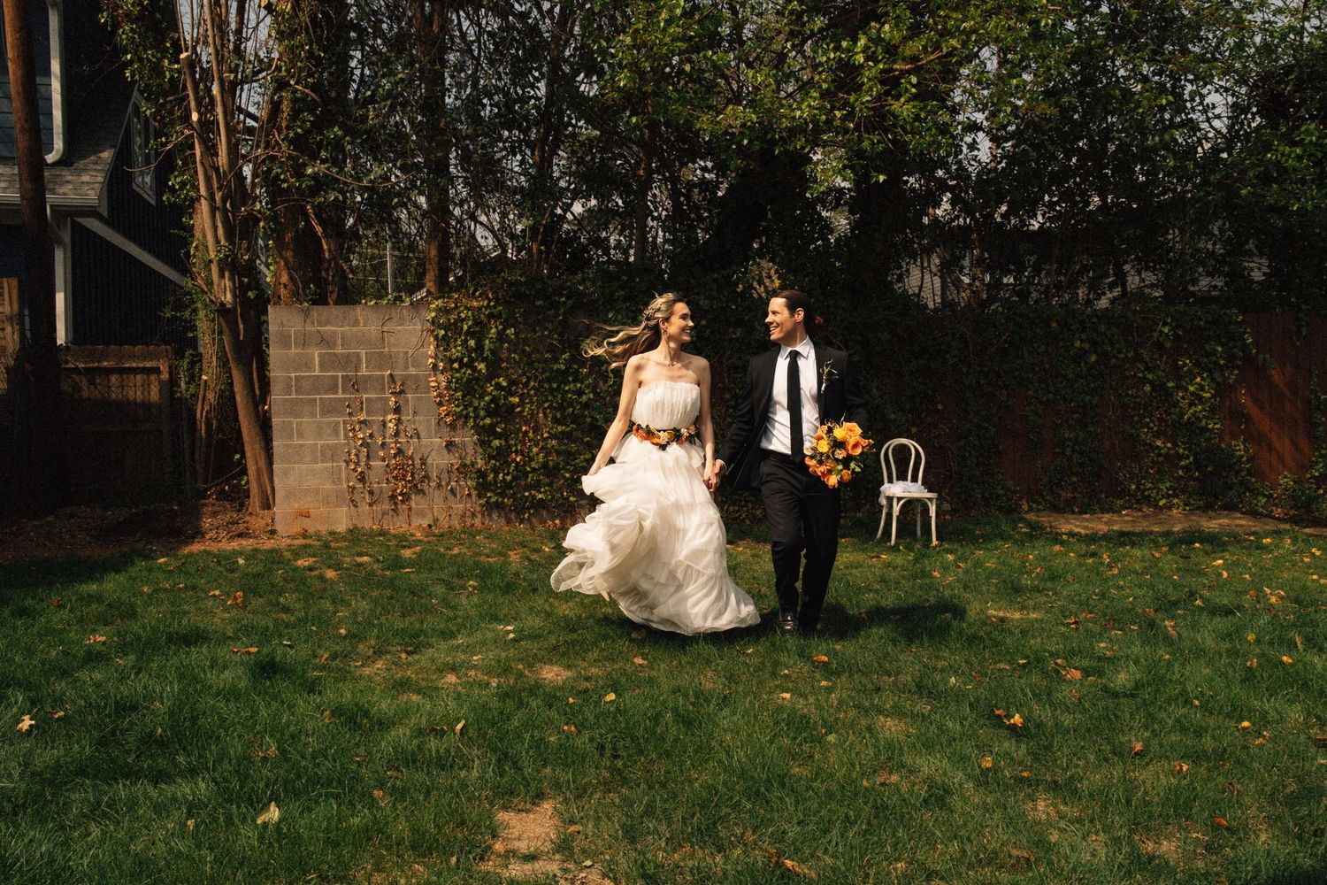 Bride and groom laughing and walking hand in hand across a lush green garden lawn on their wedding day.