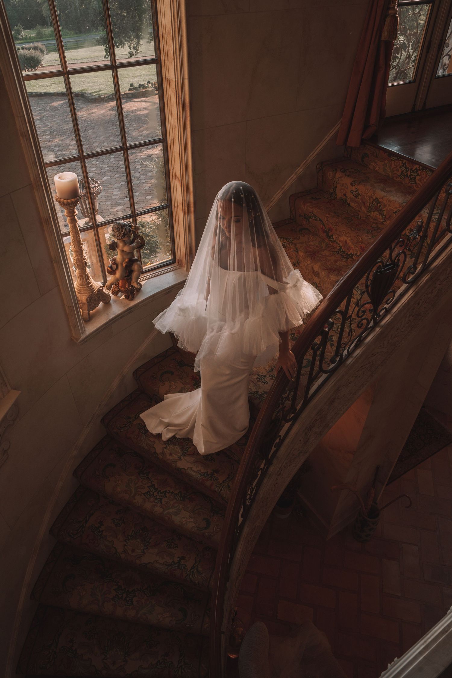 Bride in white gown and veil descending elegant curved staircase near large window in ornate venue.