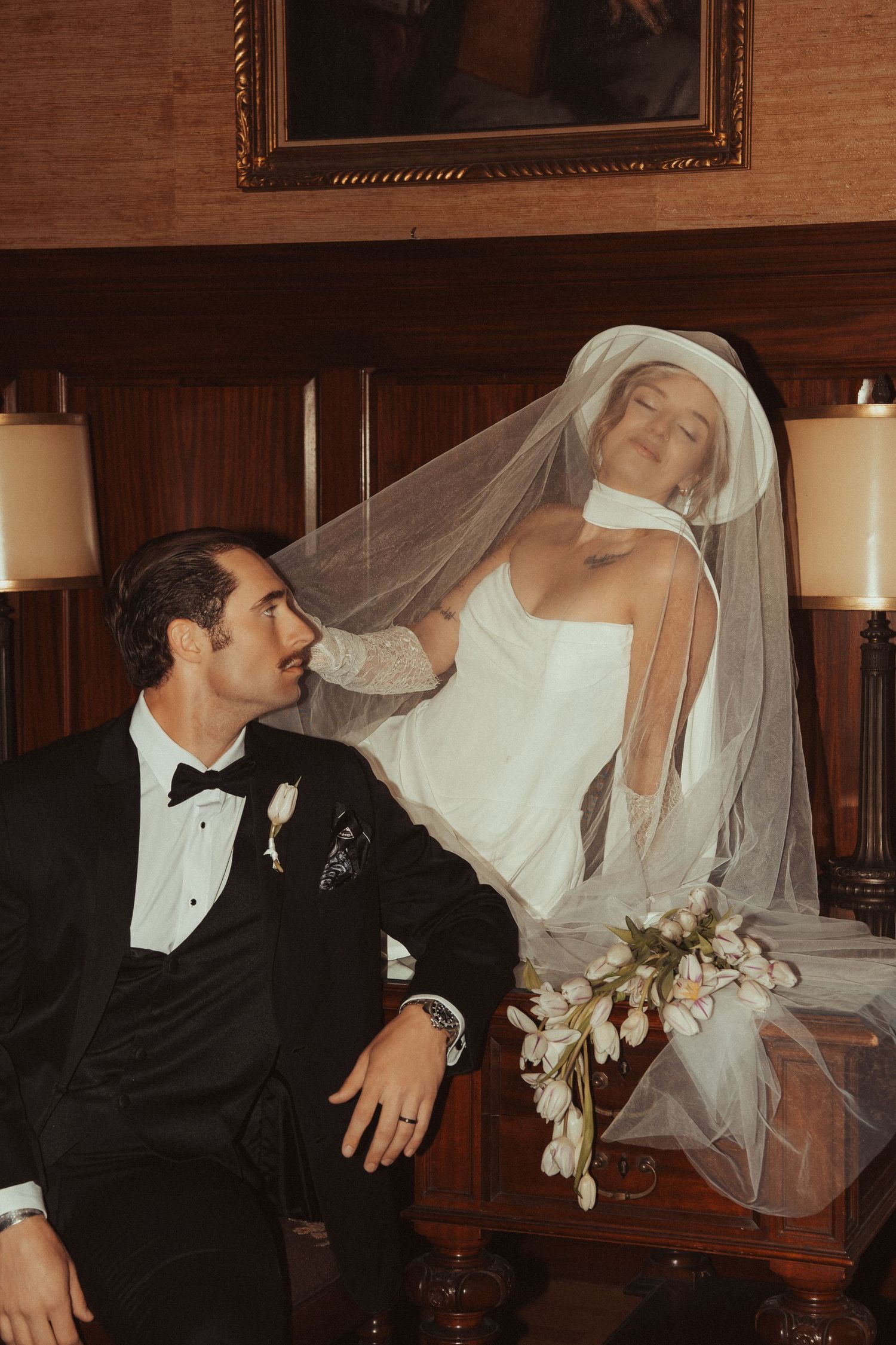 Bride in white gown and veil poses with groom in black tuxedo at elegant wood-paneled wedding venue.