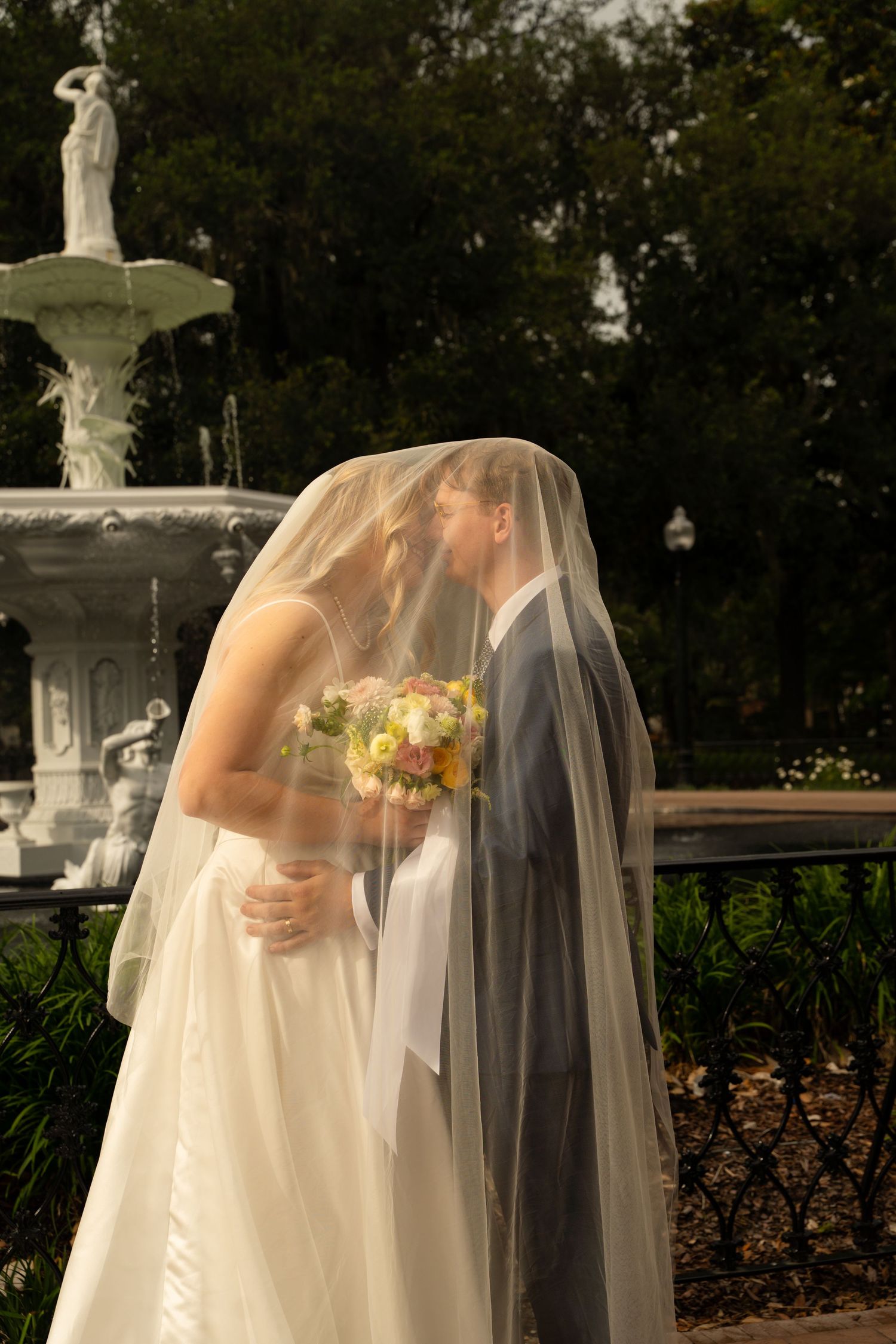 Bride and groom kissing under veil near white fountain in garden during outdoor wedding ceremony.