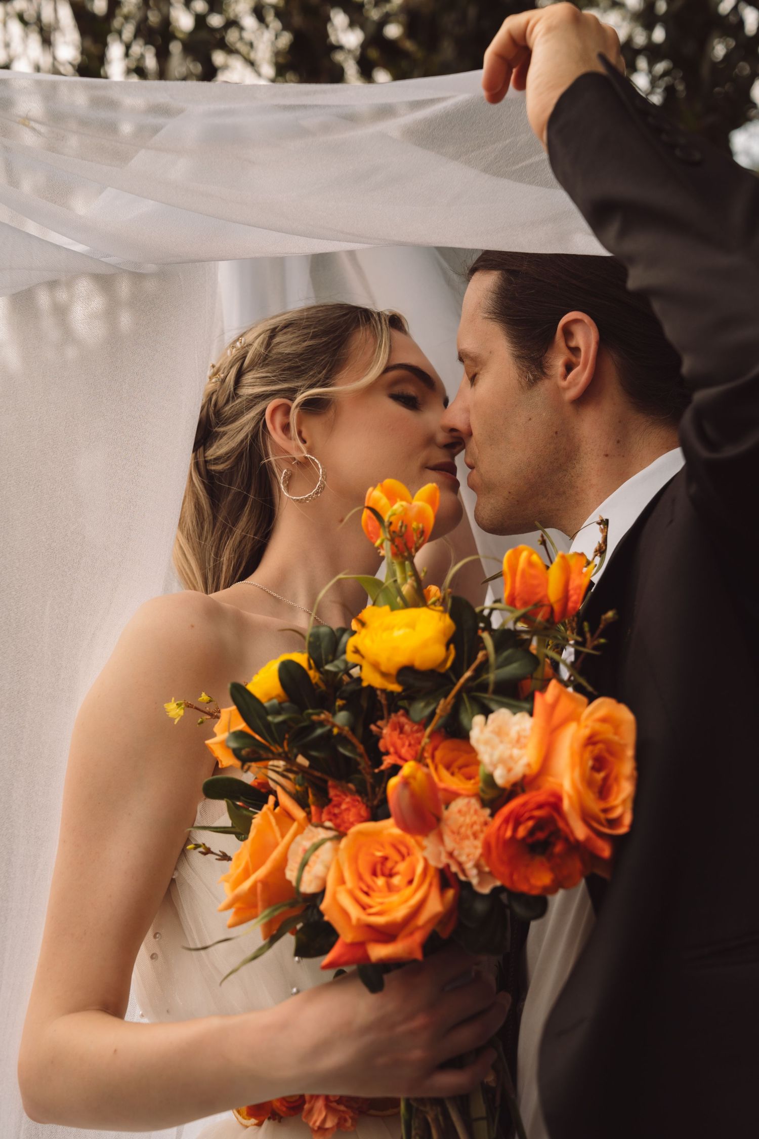 Bride and groom share a kiss under a veil holding a vibrant orange and yellow rose bouquet.