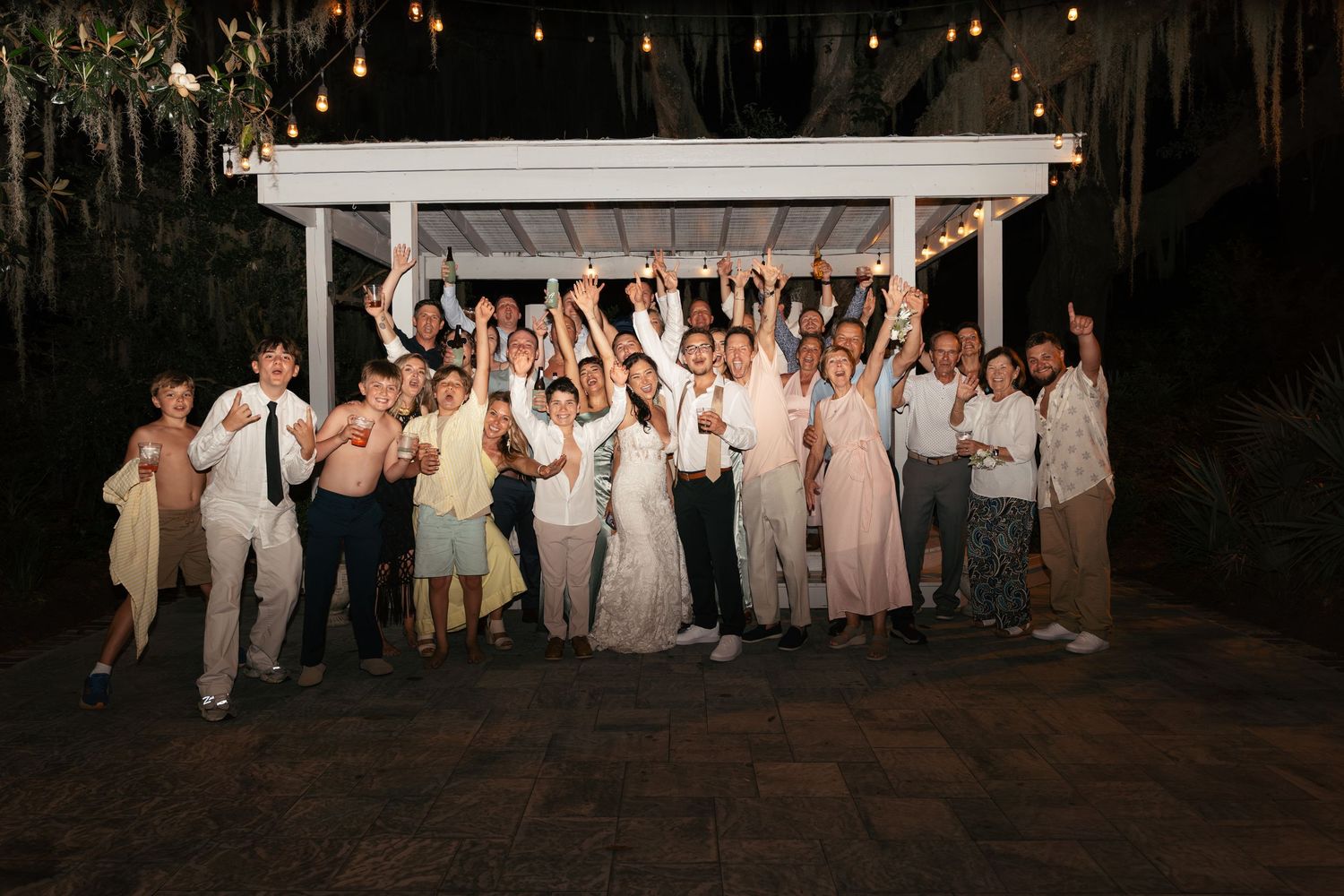 Large group of wedding guests celebrating outdoors at night under string lights and a white pergola.