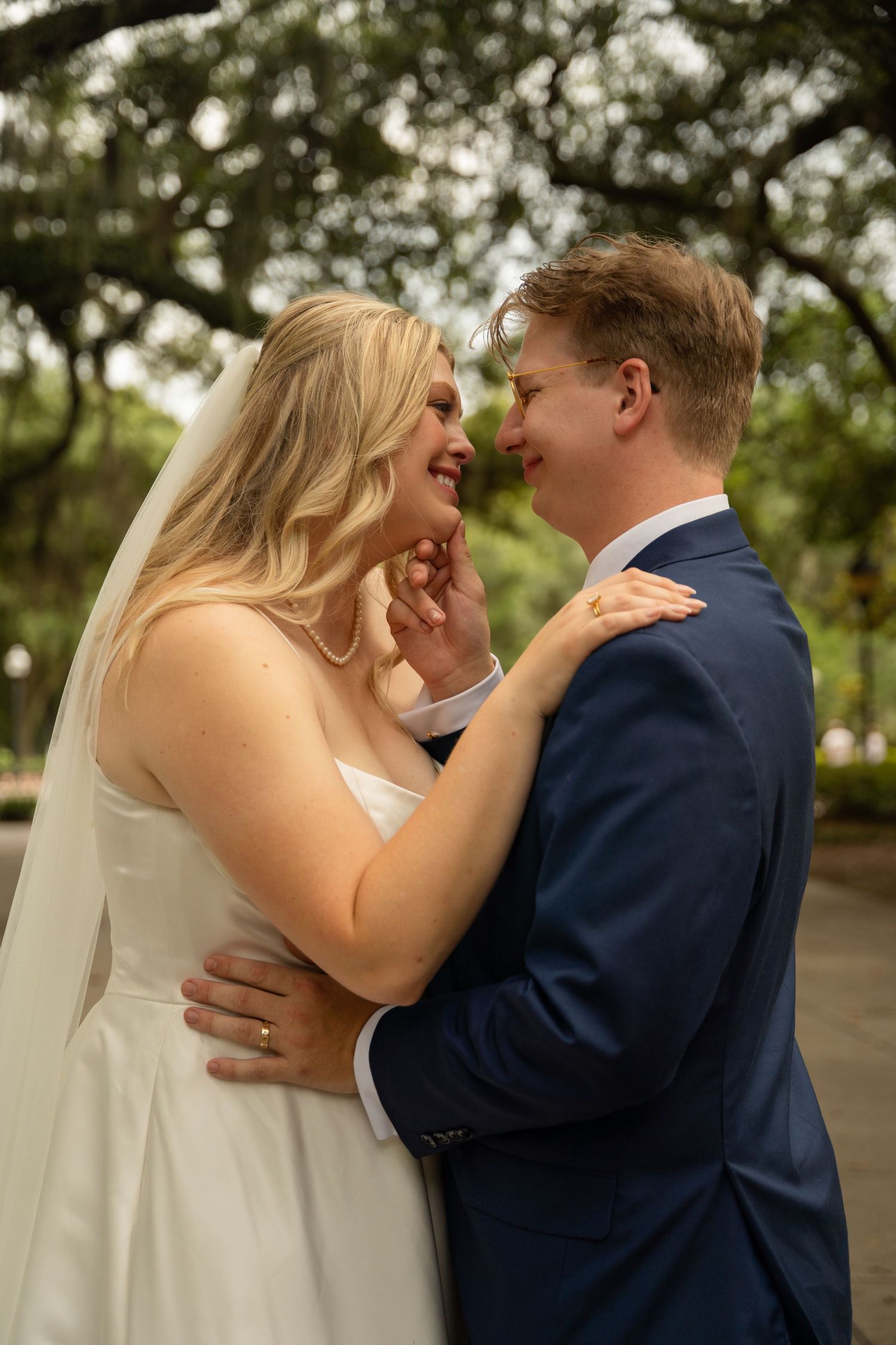 A bride and groom share a joyful moment, smiling at each other outdoors under lush green trees on their wedding day.