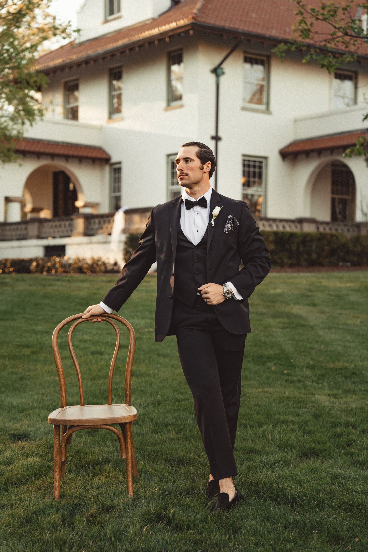 Groom in black tuxedo with bow tie stands beside wooden chair on lawn outside elegant white Spanish-style estate.