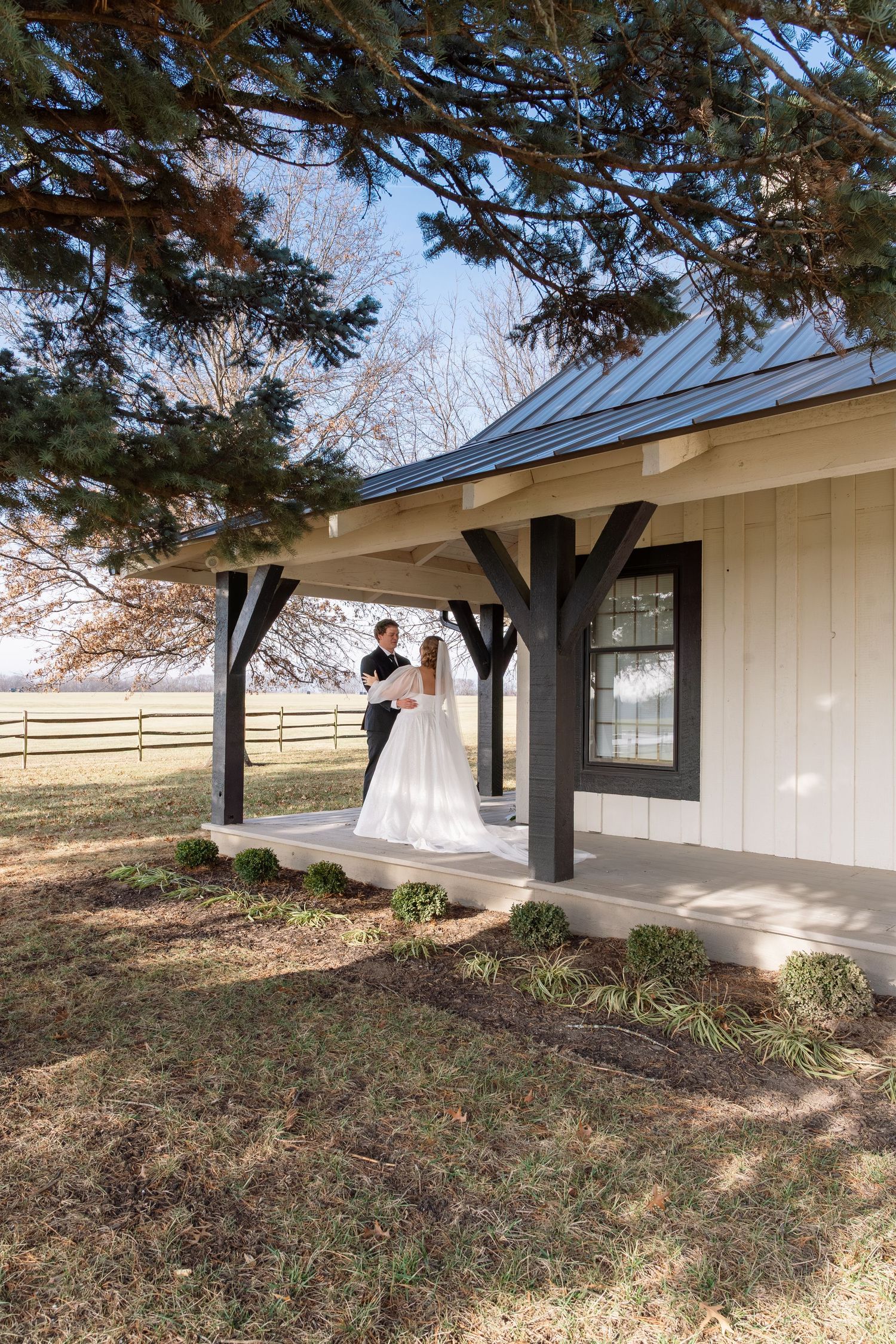 Bride and groom share tender moment on farmhouse porch under pine tree.