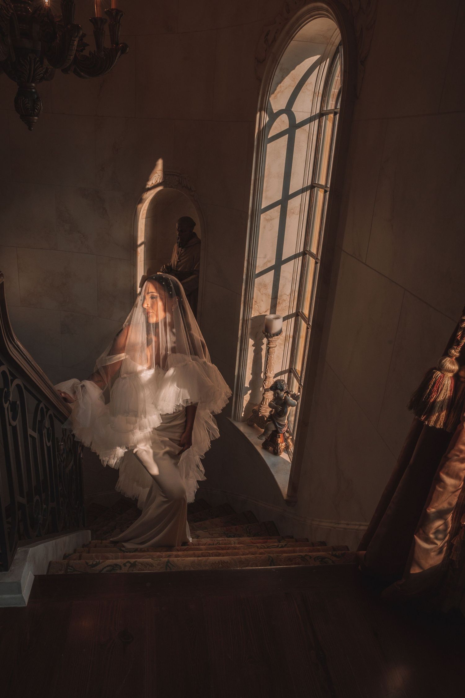 Bride in white veil and gown standing on ornate staircase bathed in golden light from arched window.