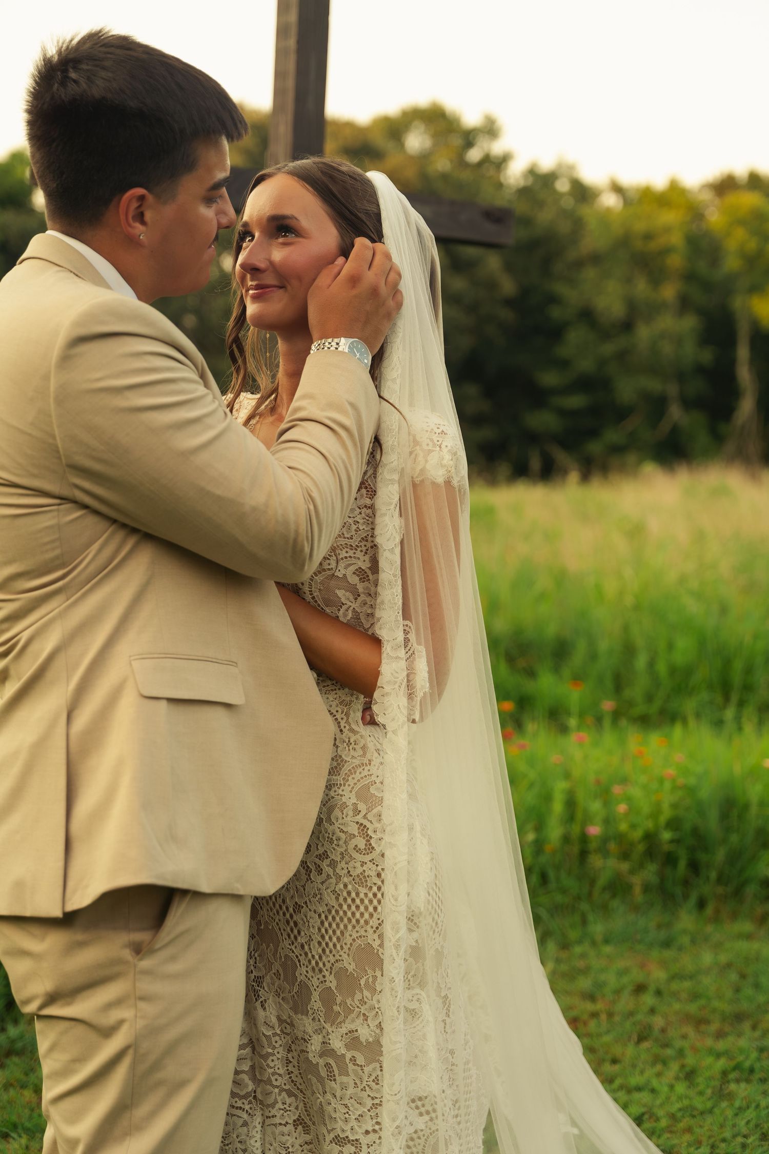 Groom in tan suit tenderly touches bride's veil outdoors, as she smiles up at him in lace wedding dress.