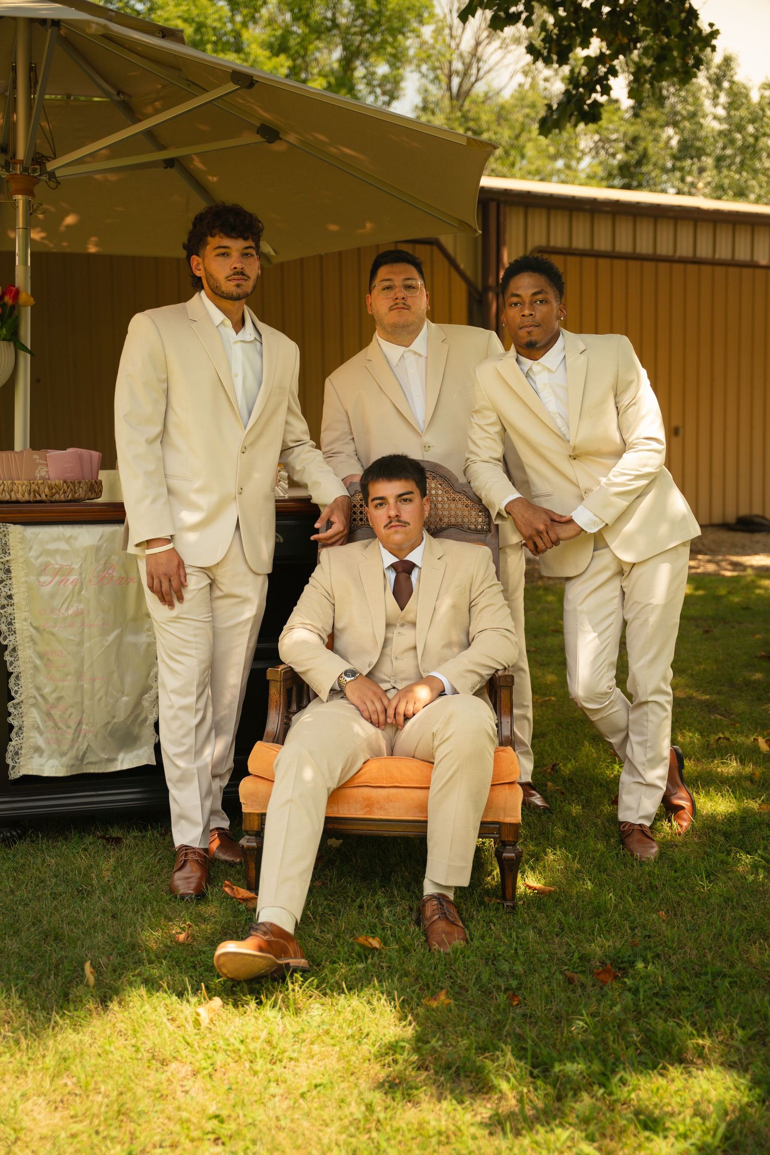 Four young men in matching cream suits pose outdoors, three standing behind one seated in a wooden chair on green grass.