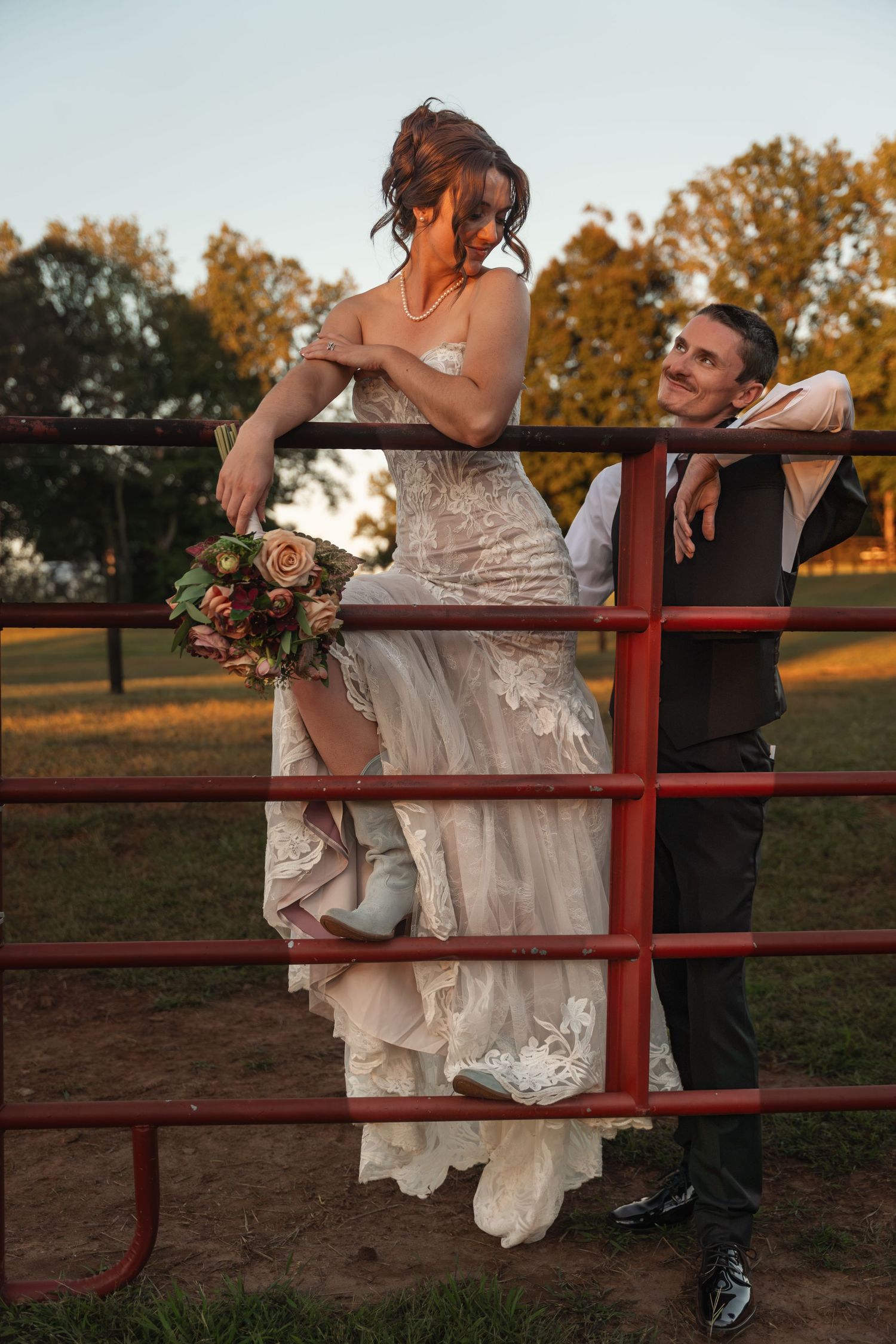 Bride in lace gown sits on red farm gate holding bouquet while groom in suit gazes up at her during golden hour.
