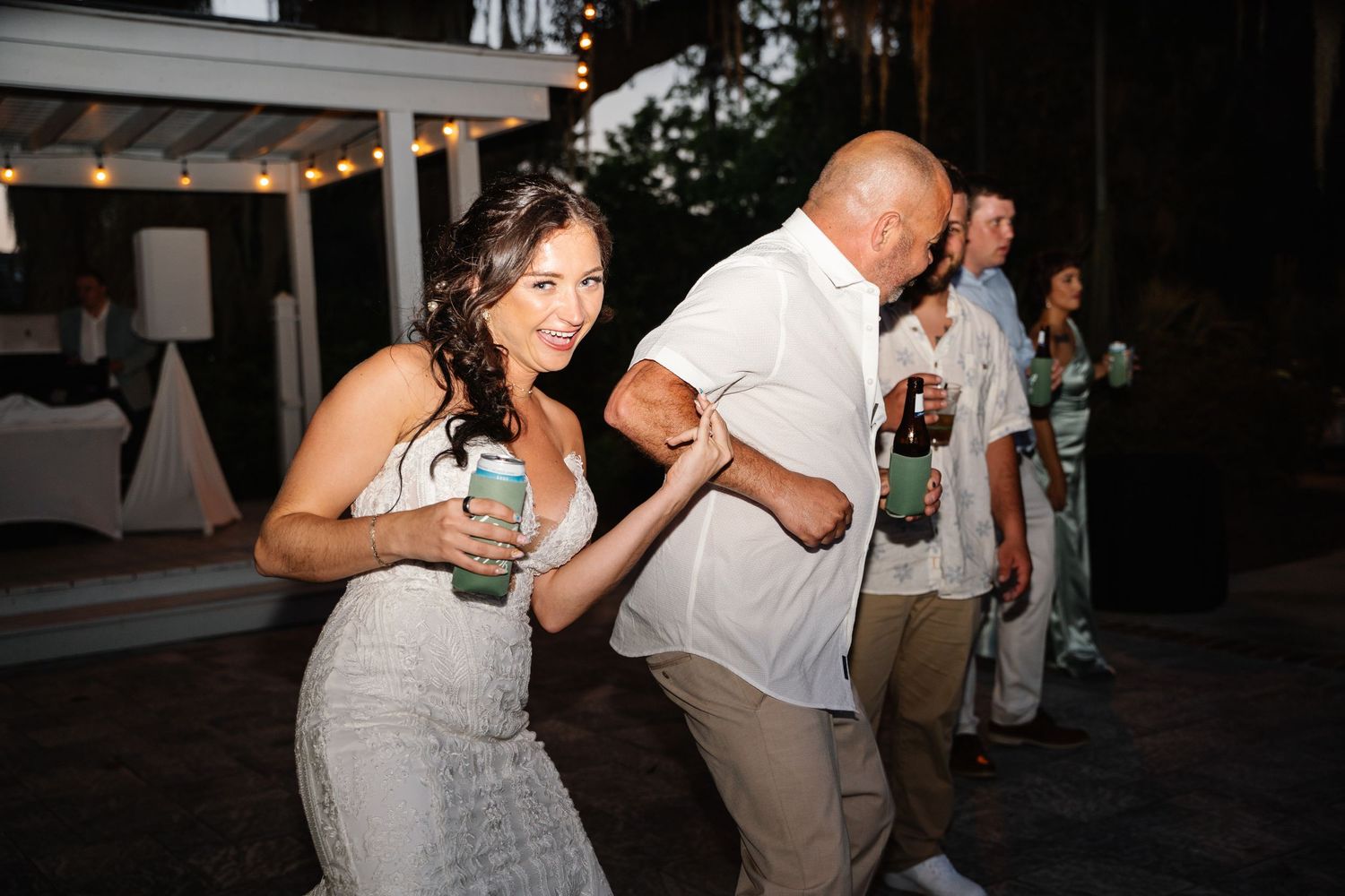 Couple dancing outdoors at a wedding reception, holding drinks and smiling under string lights at night.